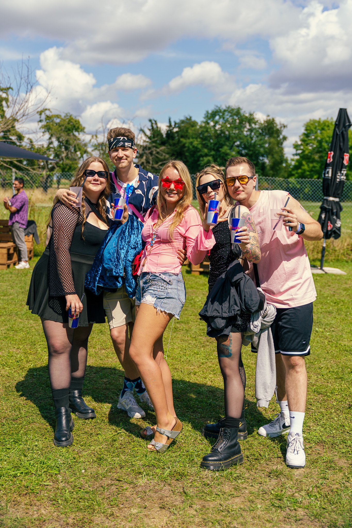 Group of five friends standing outdoors on grass, smiling and posing for a photo, holding drinks, with a few other people in the background.