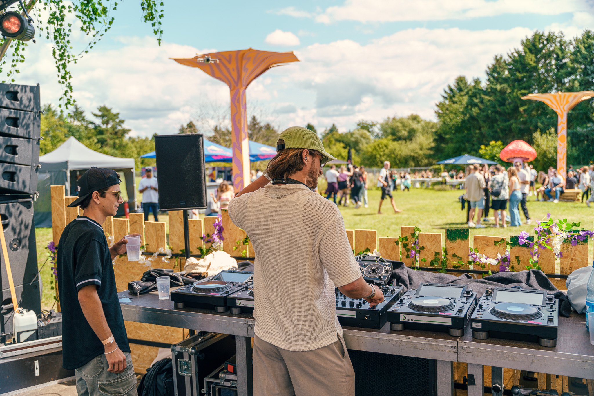 A DJ plays electronic music at an outdoor festival with people dancing on the grass. Brightly colored mushroom and tree sculptures are in the background, and tents and trees surround the scene.