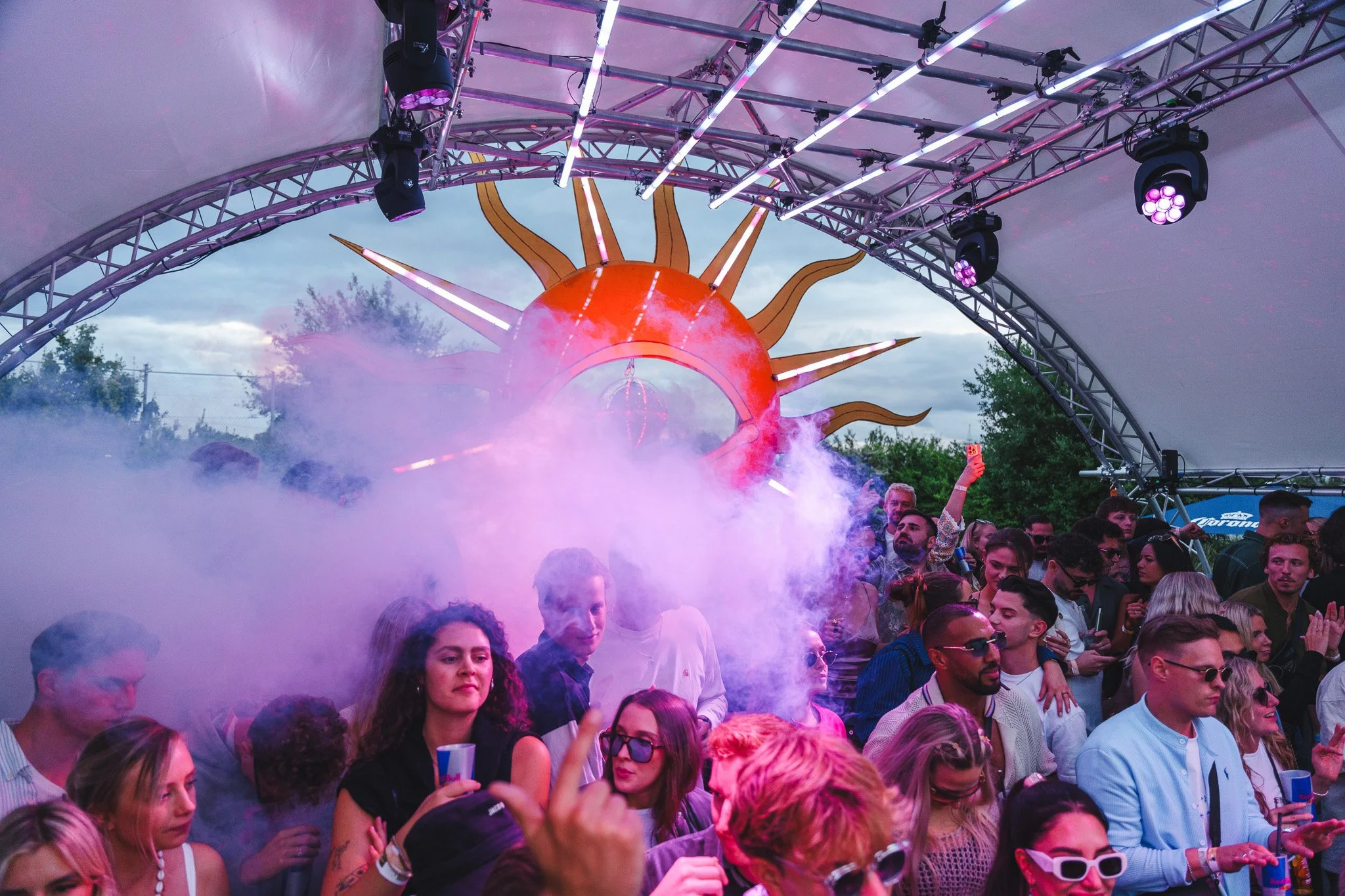 A crowd of people dancing at an outdoor music festival or concert under a large stage with a sun-themed decoration in the background, smoke effects, and purple stage lights.