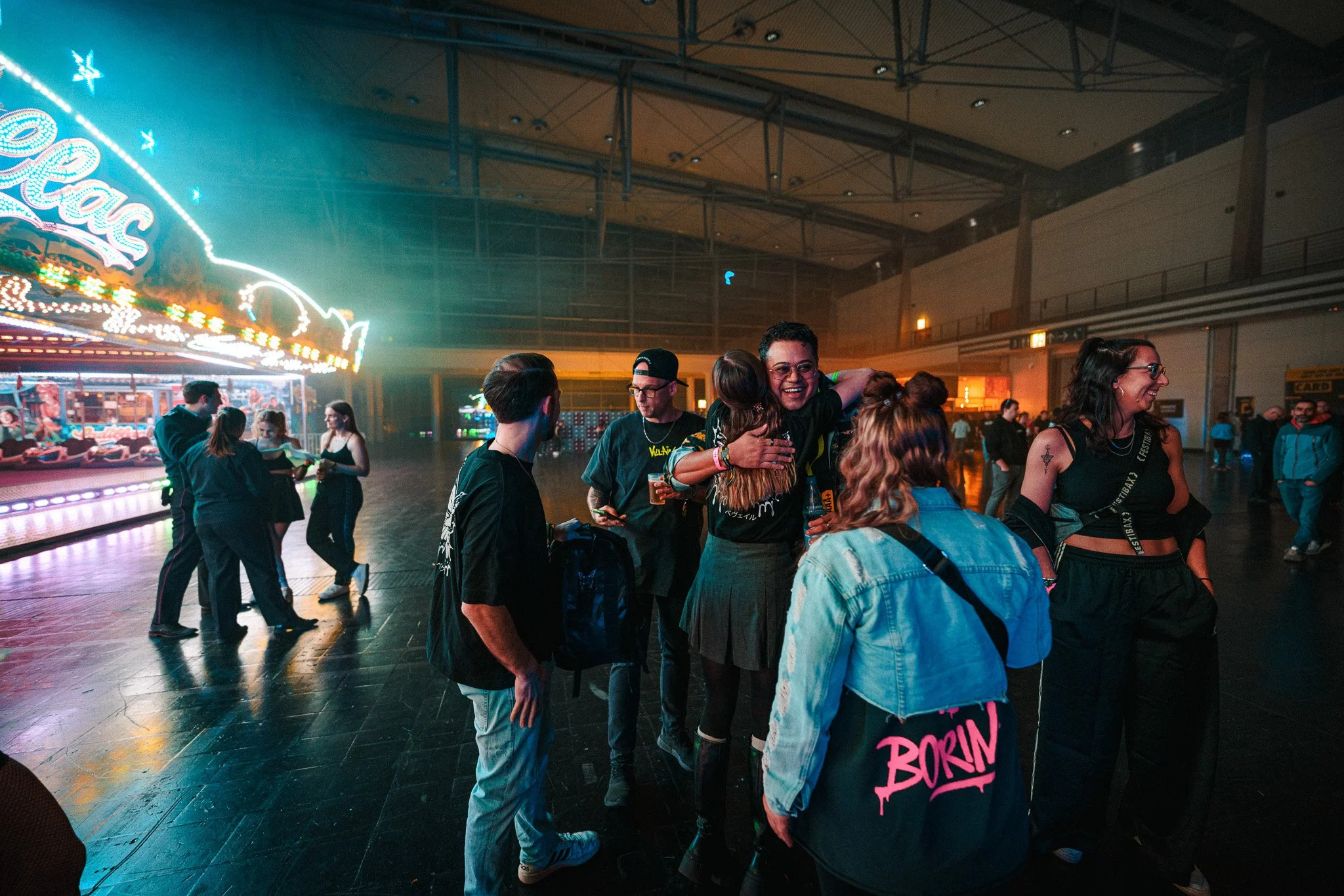 People dancing and socializing in an indoor venue with neon lights and carnival rides in the background.