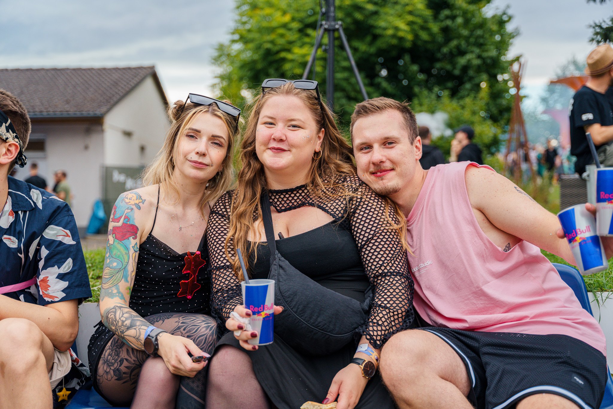 Three friends at an outdoor event sitting close together, with festival drinks in hand, smiling. The background features other people, greenery, and a cloudy sky.