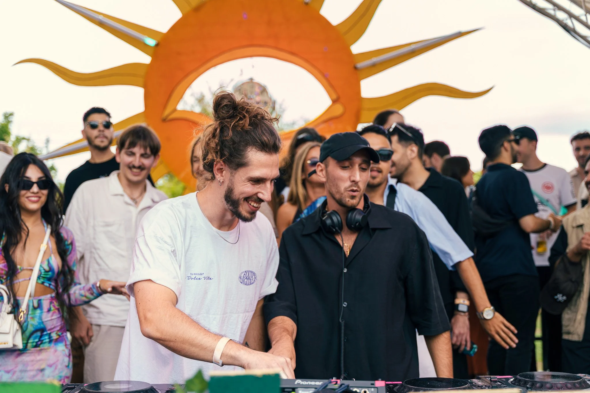 Crowd of people at an outdoor music event, with a DJ playing music in the foreground, large decorated sun sculpture in the background.