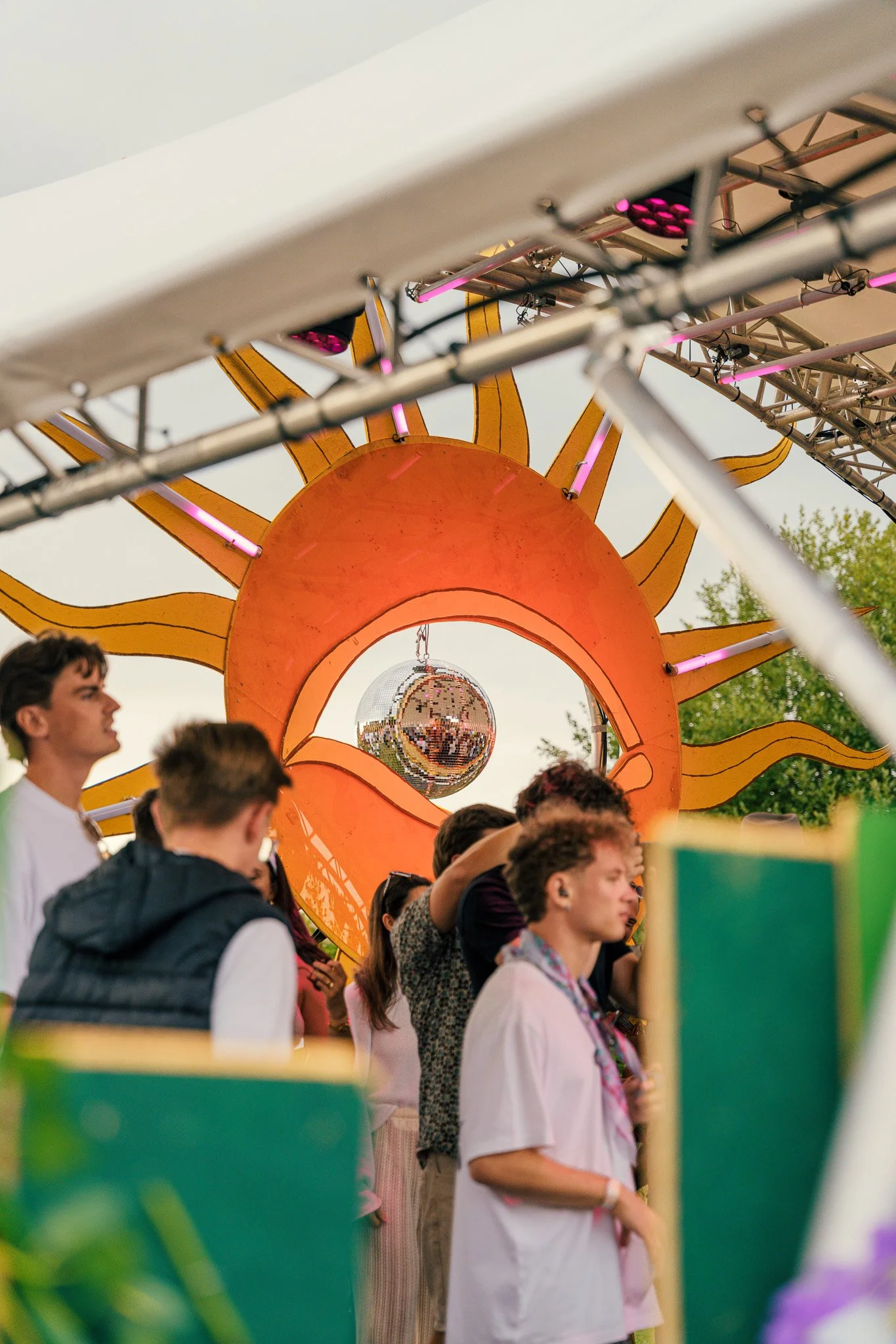 Group of young people at a music festival with a colorful sun-shaped decoration and a disco ball overhead.