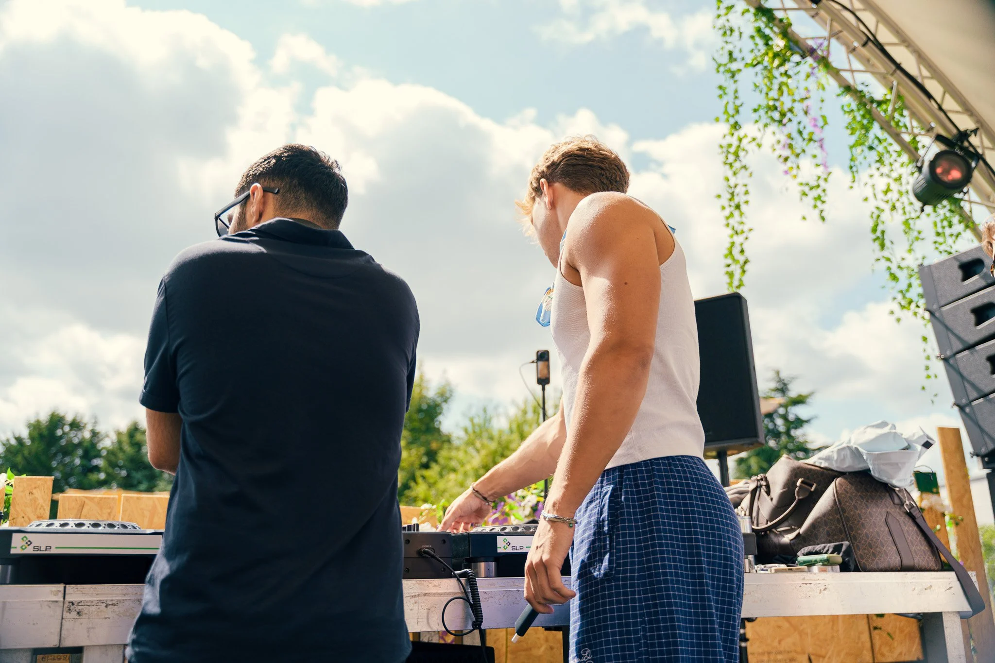 Two people are DJing outdoors with a DJ console, under a decorated tent, with trees and a cloudy sky in the background.