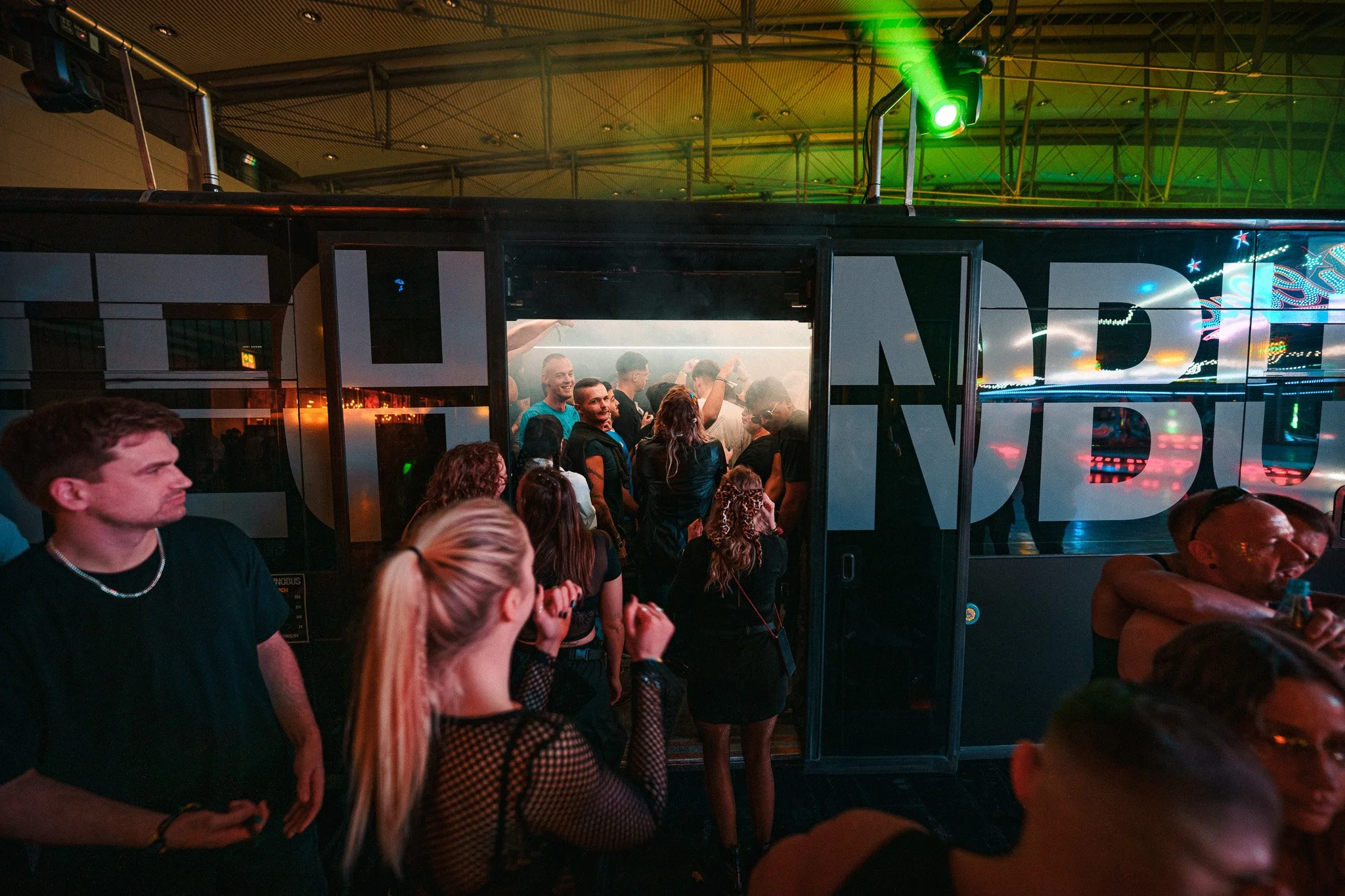 People dancing and socializing in a nightclub with colorful lights and a DJ booth.
