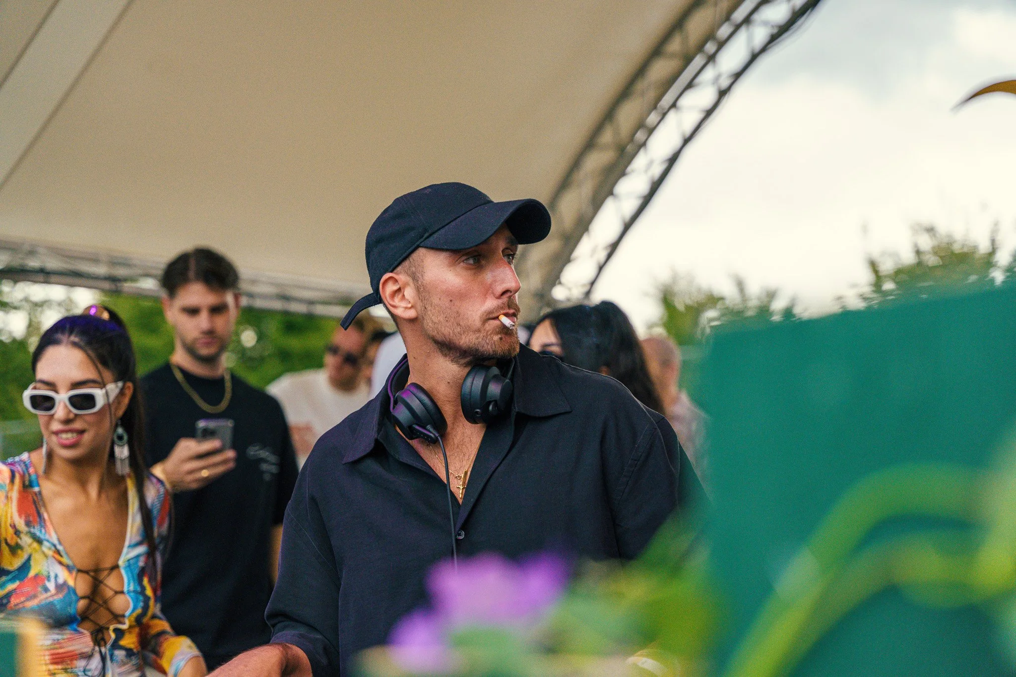 A man wearing a black baseball cap, black shirt, and headphones around his neck is standing under a tent, smoking a cigarette, with a group of people in the background, some using their phones.