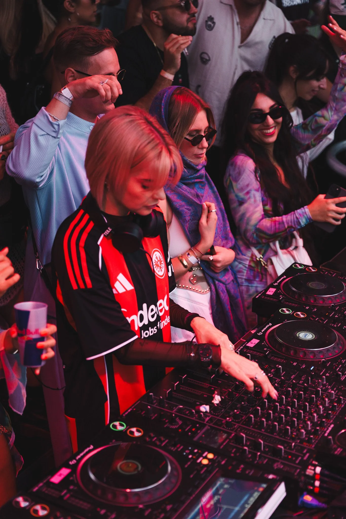 A DJ with blonde hair wearing a red and black sports jersey, headphones around his neck, playing music at a crowded event. People around him are enjoying the music, some taking photos and wearing sunglasses.