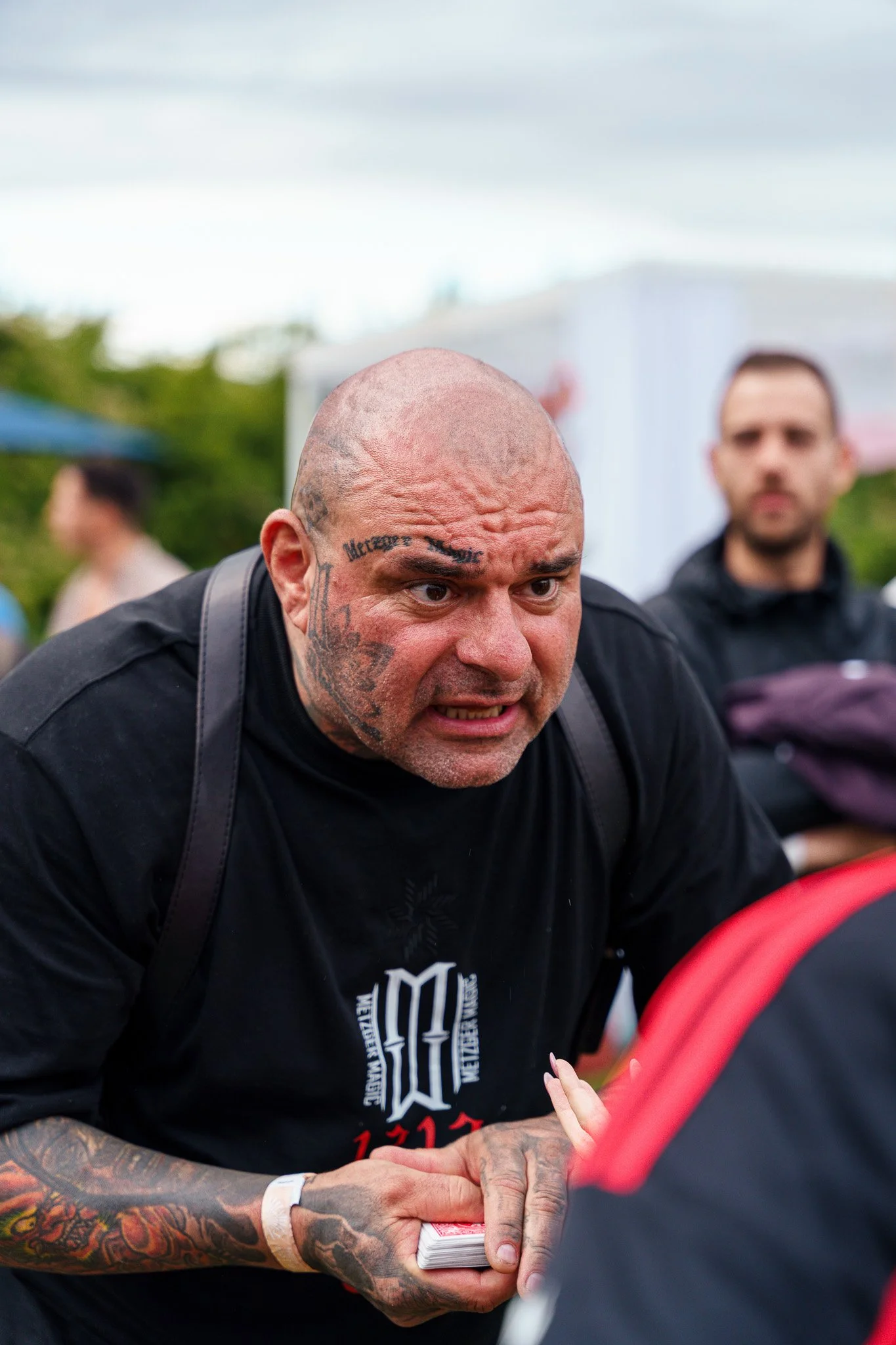 A tattooed man with a shaved head is intensely engaged in a conversation, holding a deck of cards in his hands, with blurred people and outdoor setting in the background.