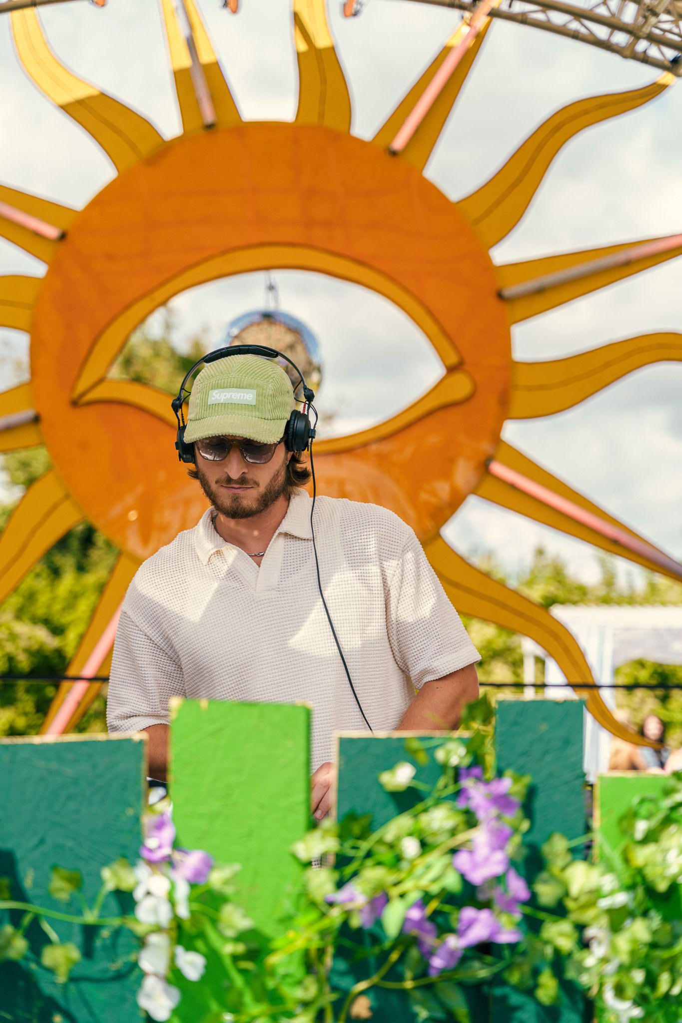 A man wearing sunglasses, a green Supreme hat, and headphones stands in front of a colorful sun-themed amusement park ride, with purple and white flowers in the foreground.