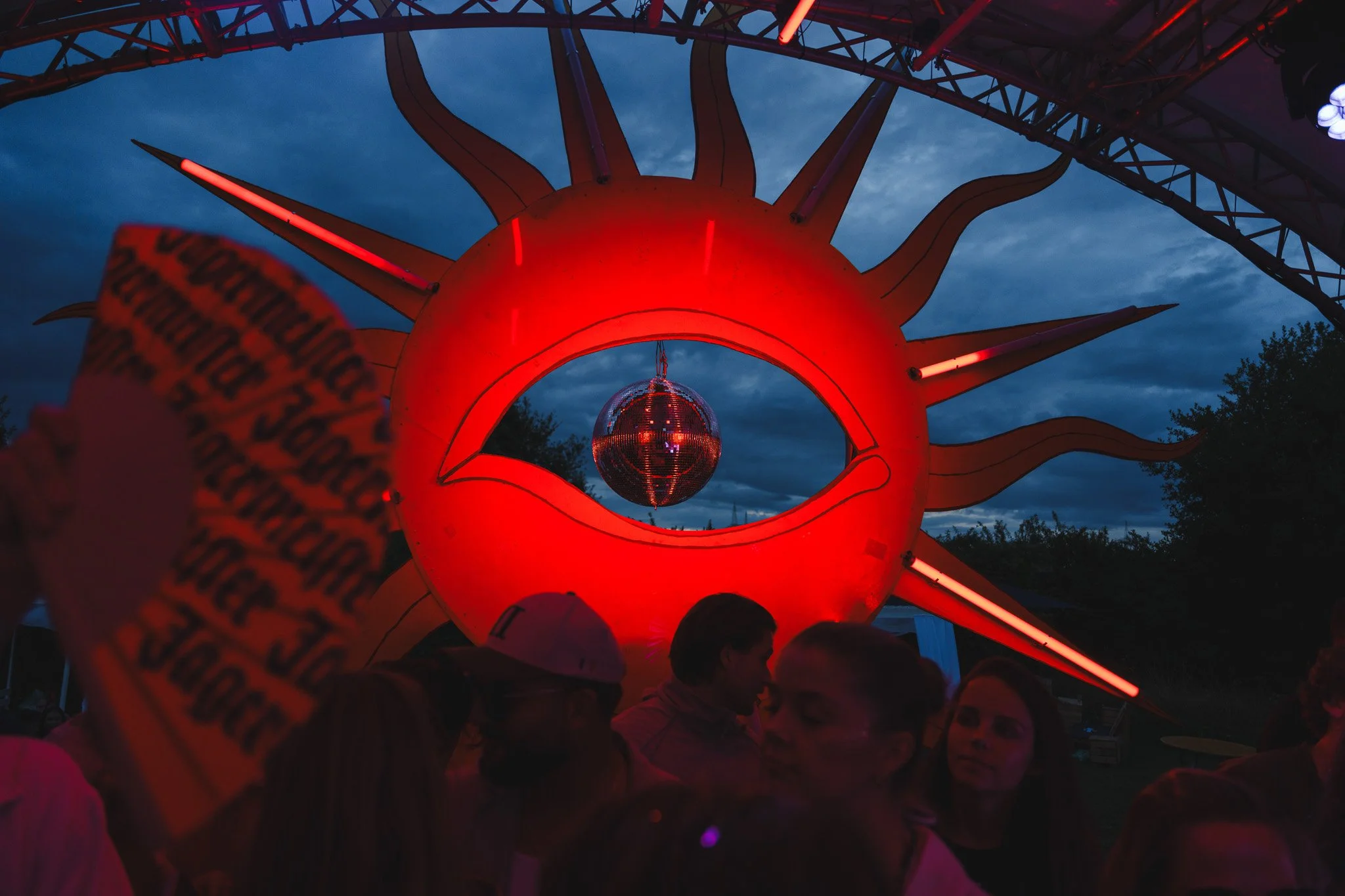A large red artistic installation shaped like a sun with an eye in the center, featuring a disco ball hanging inside the eye, illuminated during the evening with a crowd of people gathered around.
