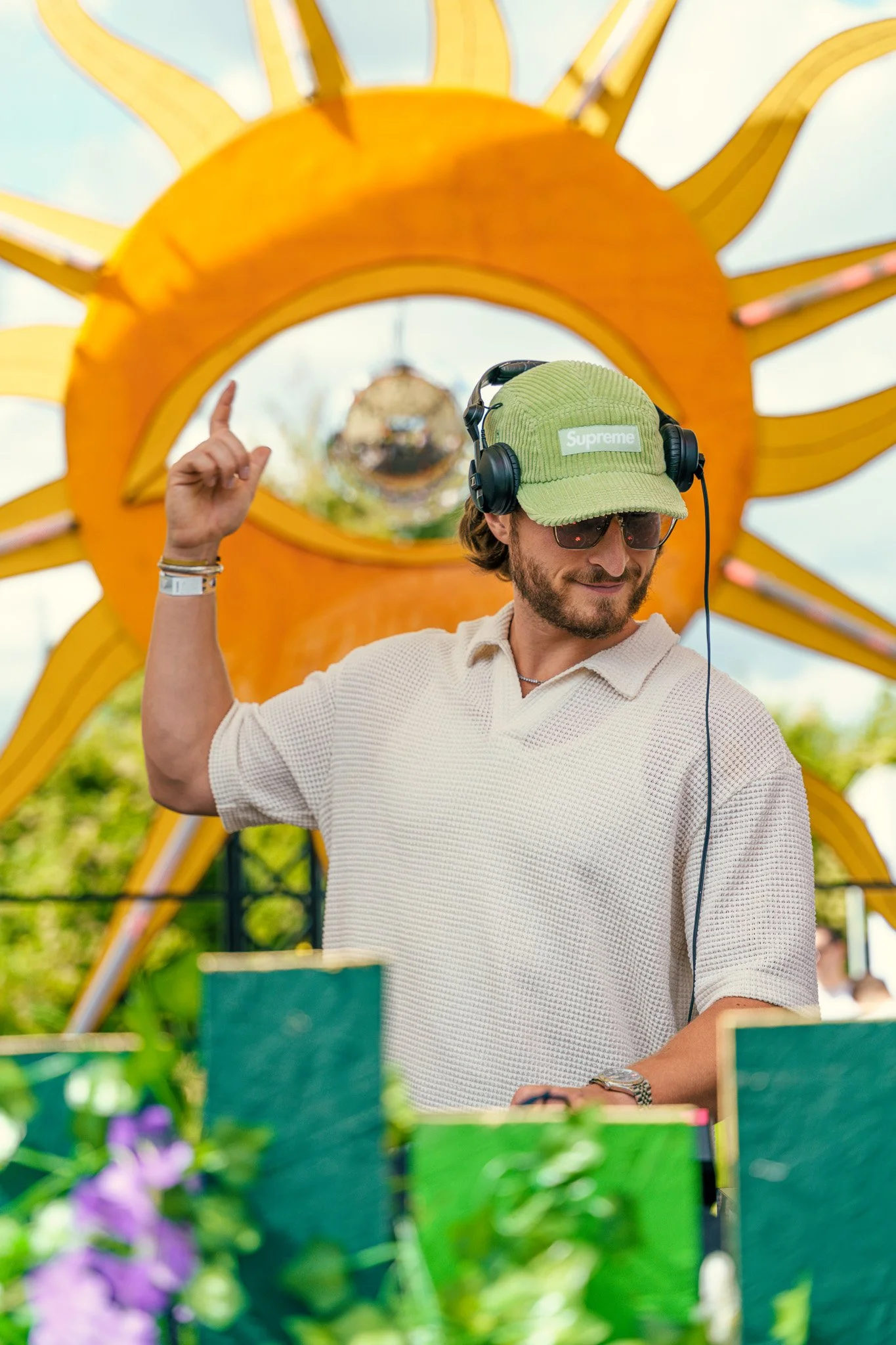 A man wearing sunglasses, a green Supreme cap, and a white polo shirt, DJing outdoors with a large sun decoration in the background.