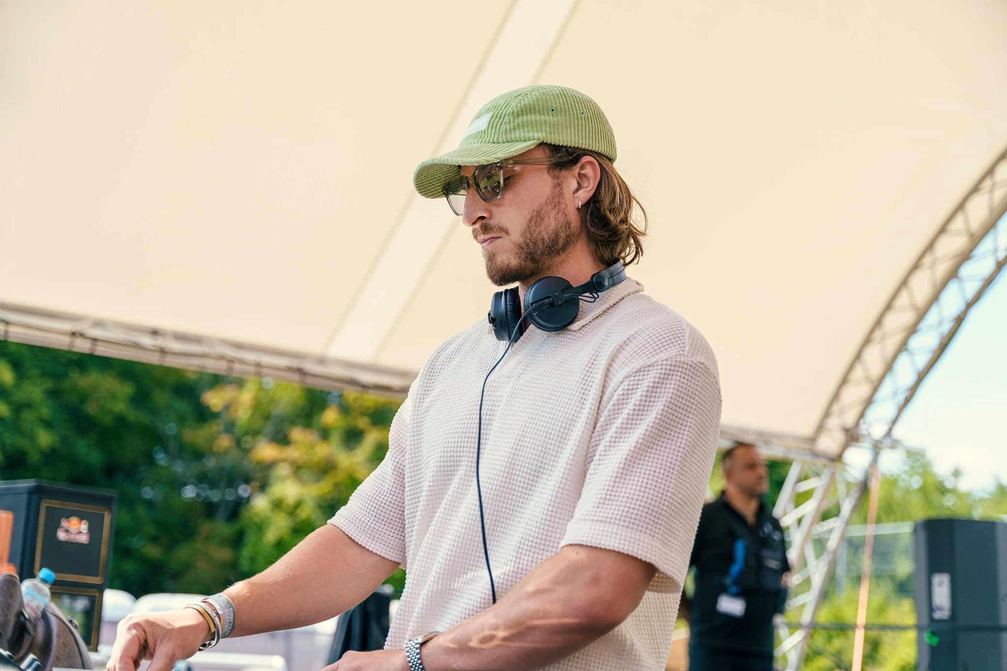 A DJ with long hair, sunglasses, and a green cap wearing headphones around his neck, performing outdoors under a white canopy.