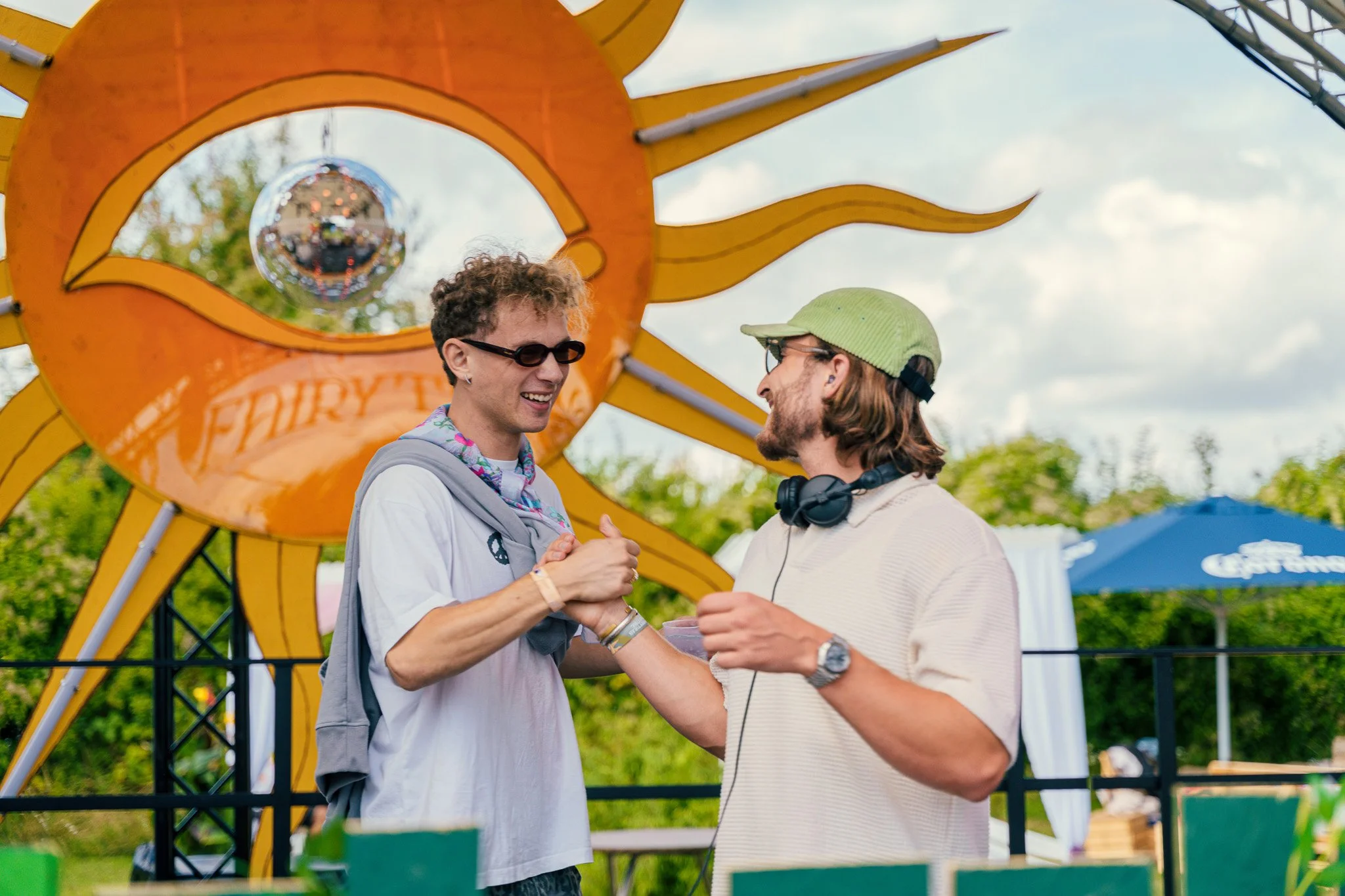 Two young men are smiling and holding hands at an outdoor festival, with a large orange fruit-shaped structure and trees in the background.