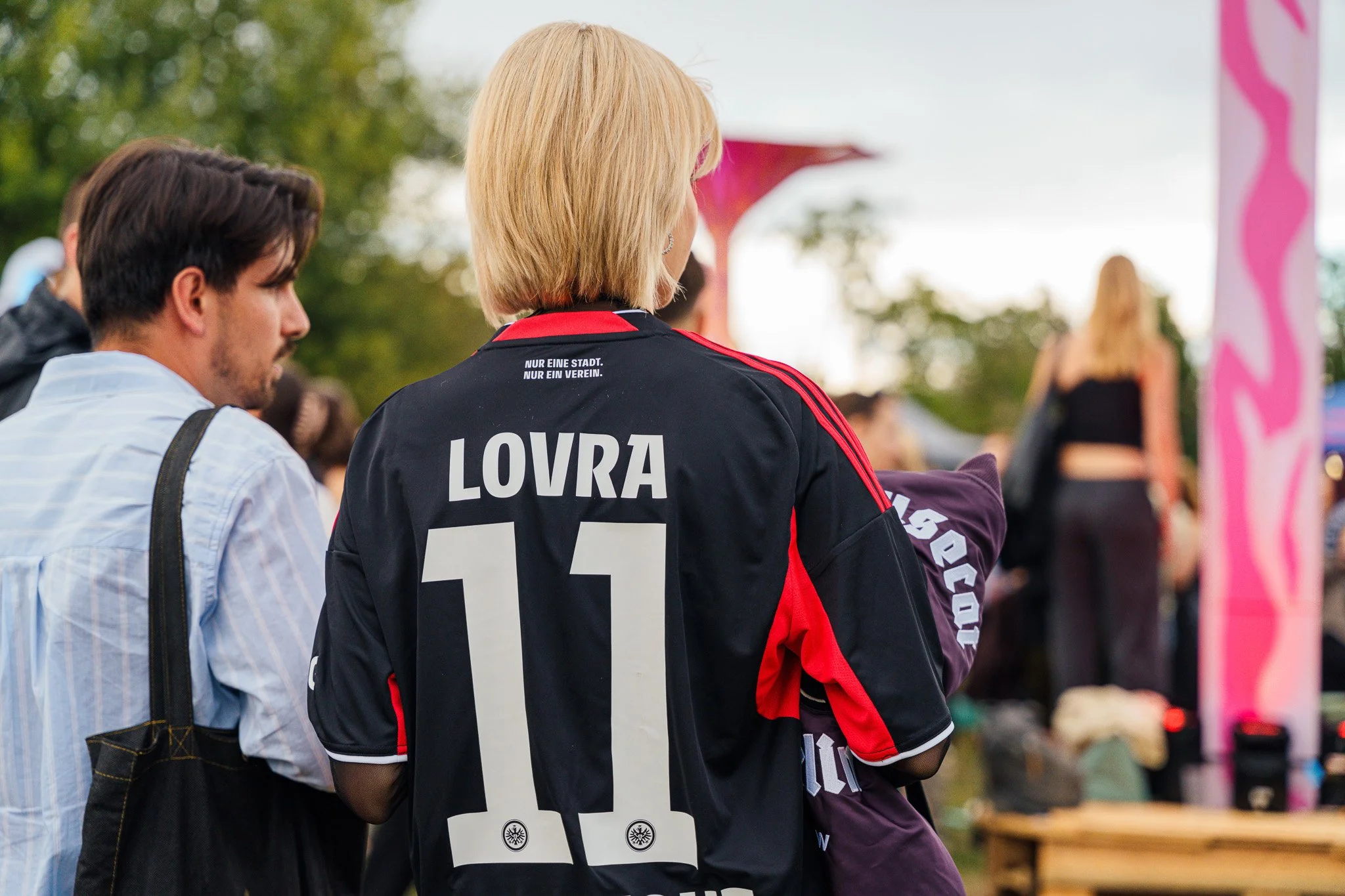 A woman with blonde hair wearing a black and red jersey with the name LOVRA and the number 11 on the back, surrounded by other people outdoors.