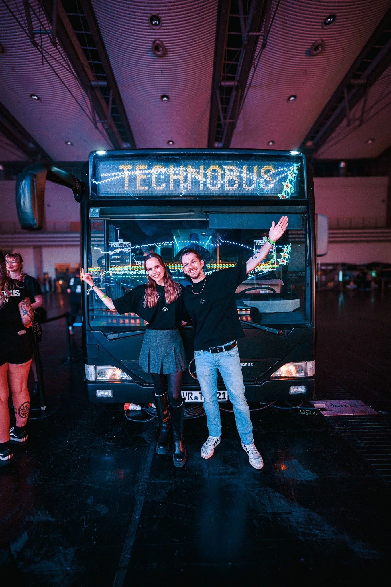 Two people posing in front of a bus with a sign that reads 'TECHNOBUS' inside a large indoor venue.