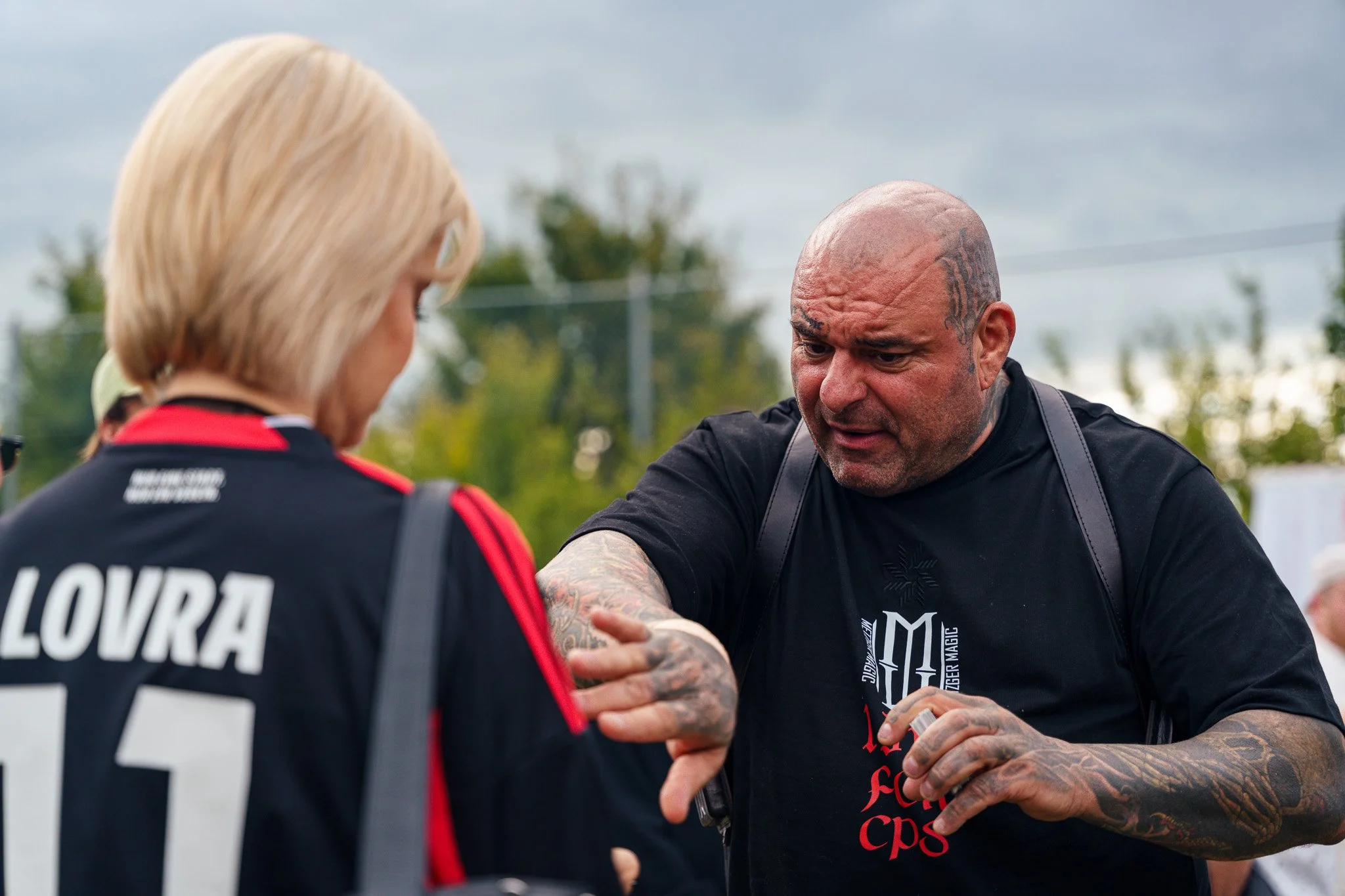A man with tattoos on his head and arms is talking to a woman wearing a black and red sports jersey with the name 'Louwra' and the number '15' on the back. They appear to be outdoors, possibly at a sports event or gathering.