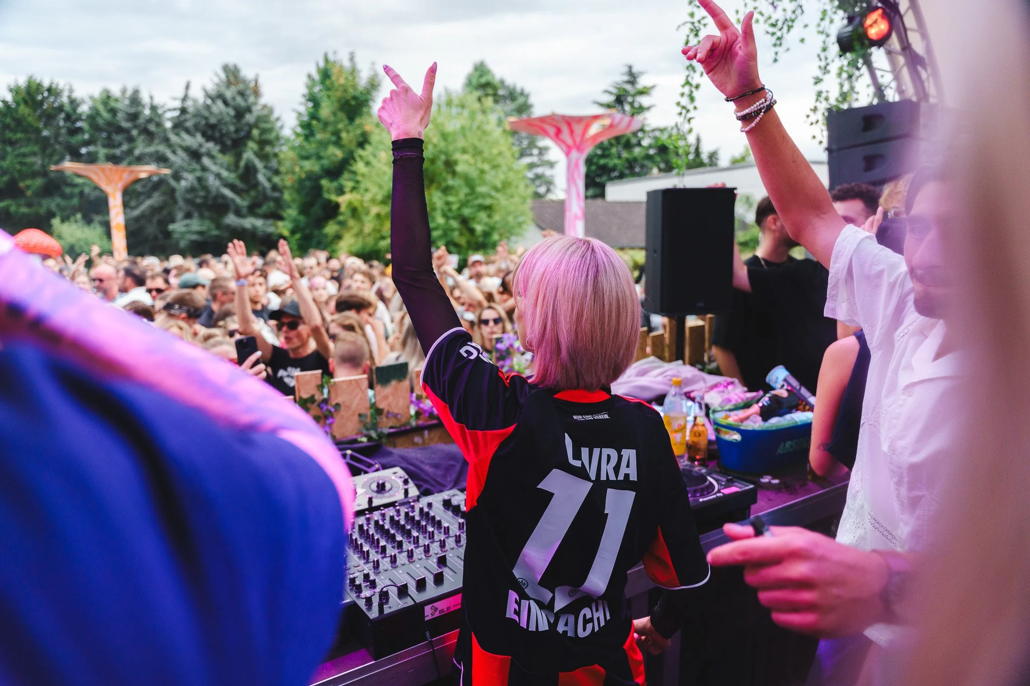 A DJ with pink hair and a black and red sports jersey is performing at an outdoor event, surrounded by a crowd of people dancing and enjoying the music during daytime.