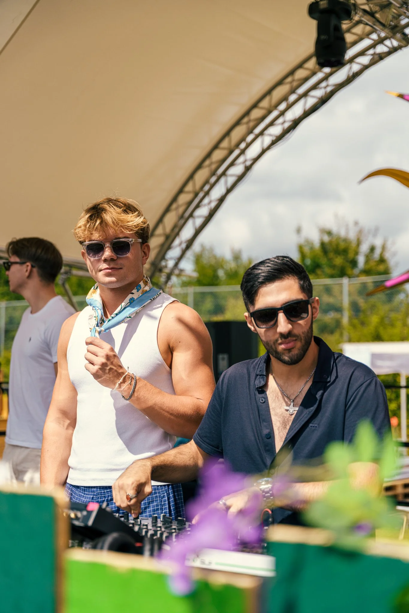 Two men with sunglasses at an outdoor event, one DJ with headphones and equipment, another casually dressed man, with a blurred background and colorful decorations.
