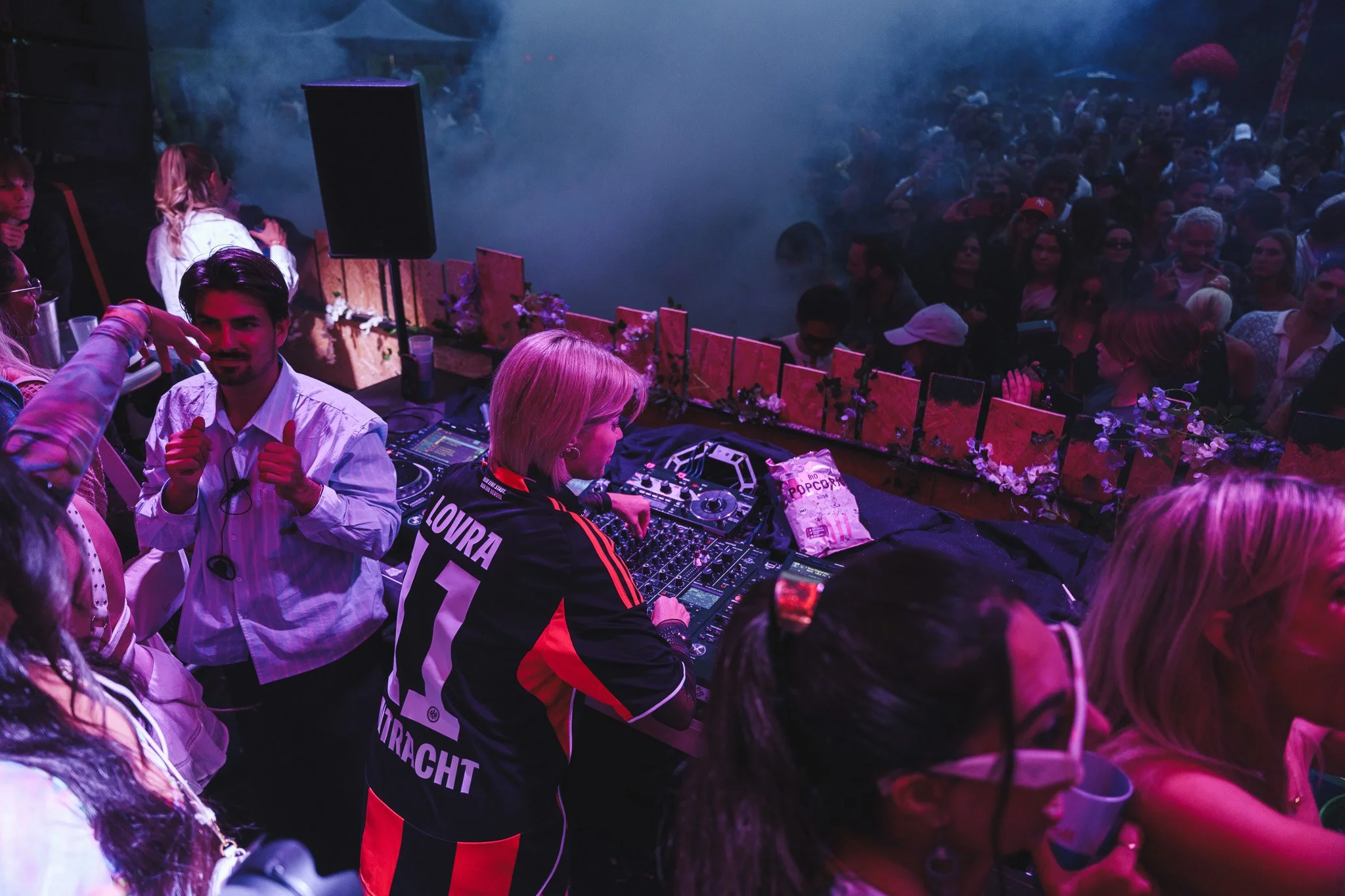 D.J. playing music at a crowded outdoor party or festival during nighttime with purple lighting, smoke, and people dancing in the background.