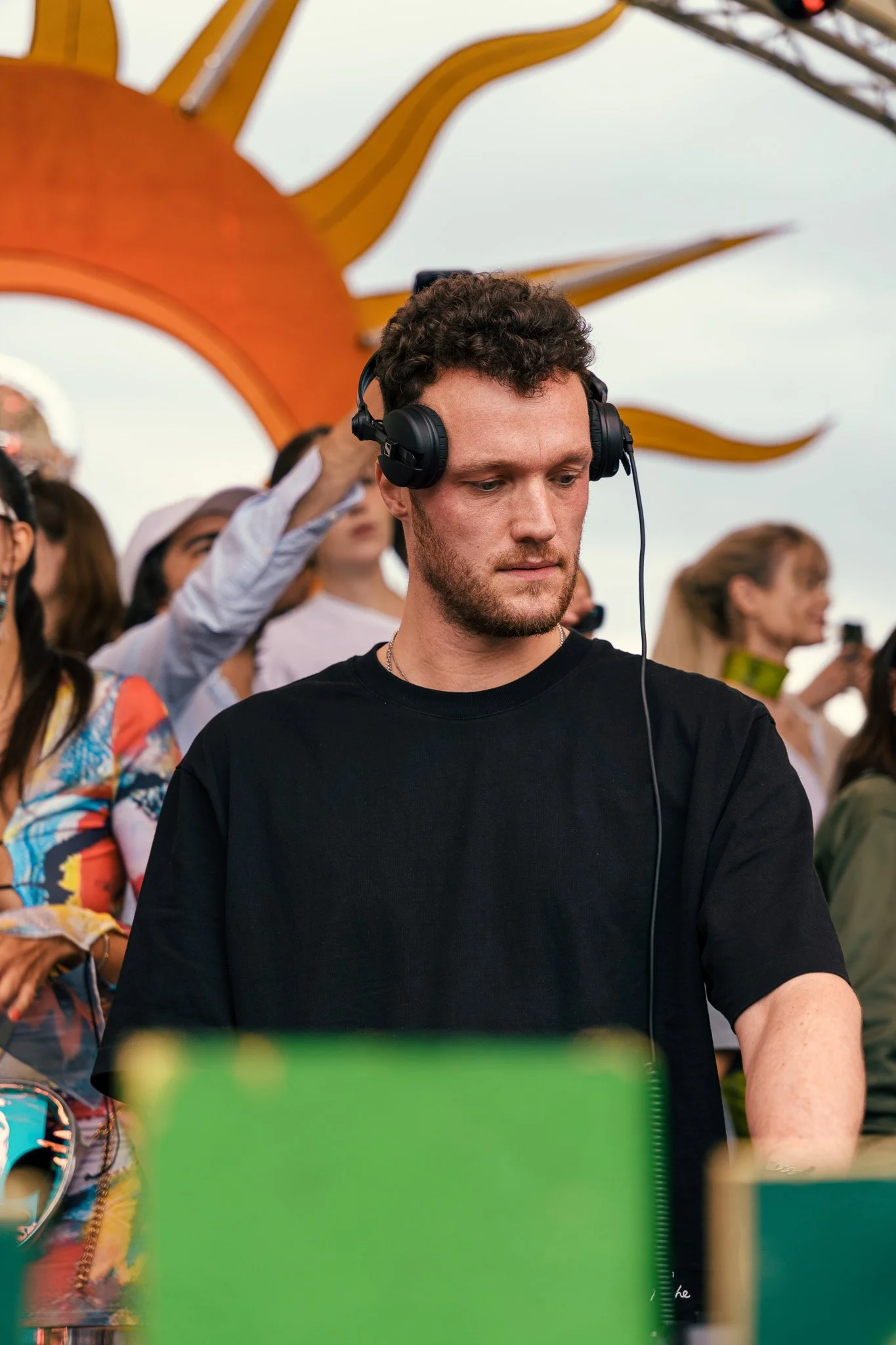 A young man with curly hair and a beard wearing a black T-shirt and headphones, standing outdoors during daytime at a public event, with a crowd of people behind him and a decorative sun-like structure in the background.