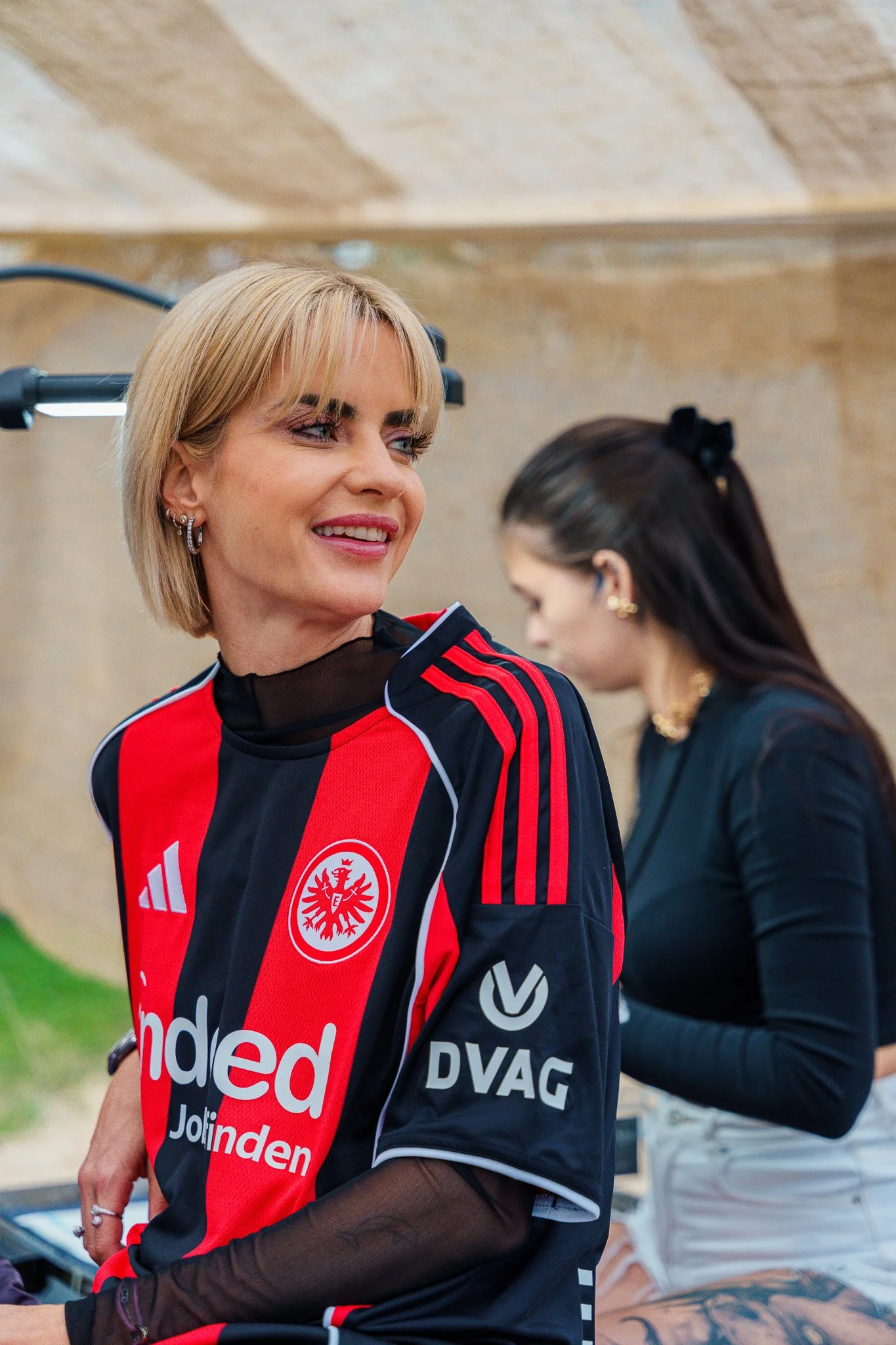 Woman wearing a black and red soccer jersey with logos and sponsor names, smiling, with a woman in black in the background.