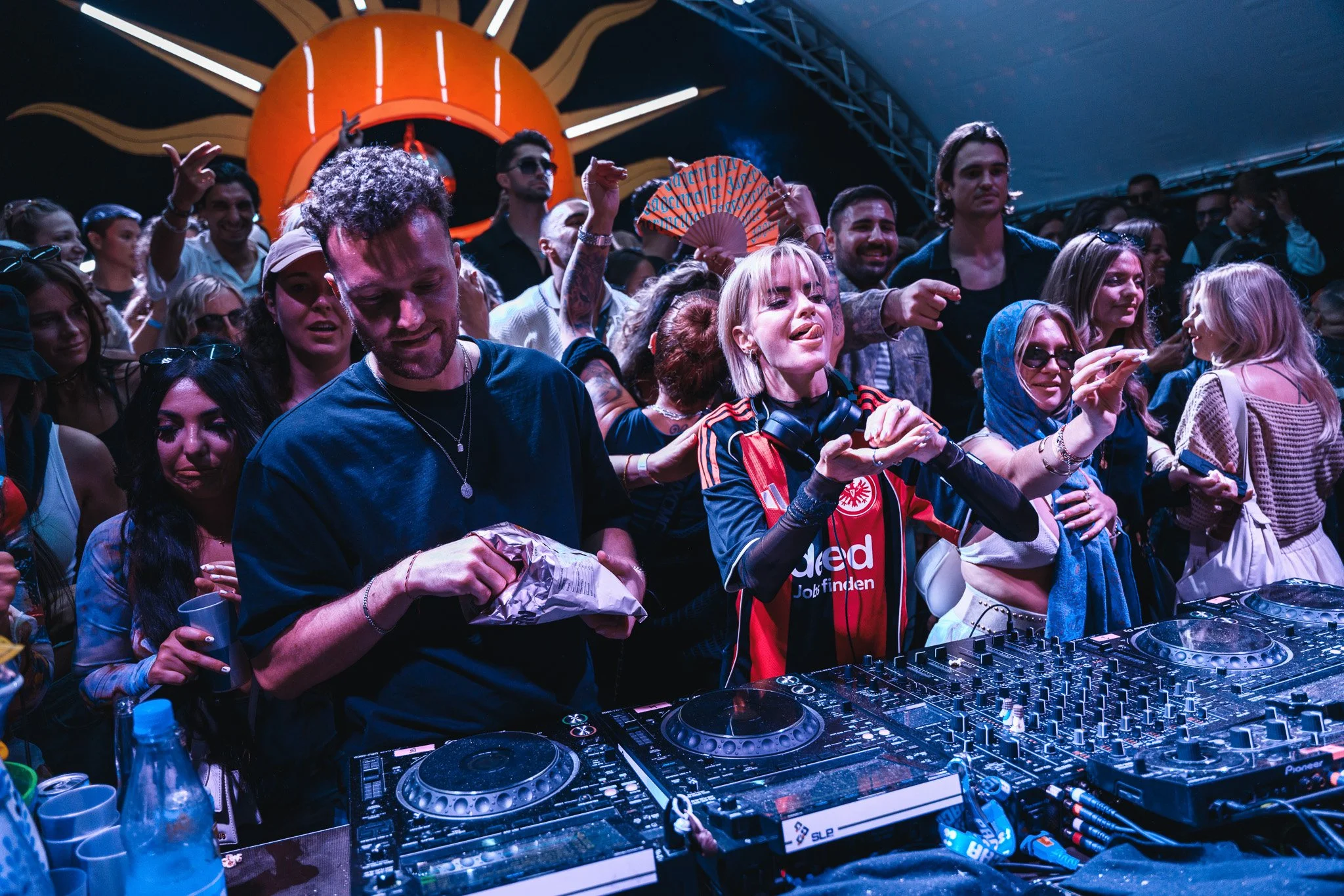 A DJ performs at a crowded music event with a large sun decoration in the background, surrounded by enthusiastic people dancing and enjoying the music.