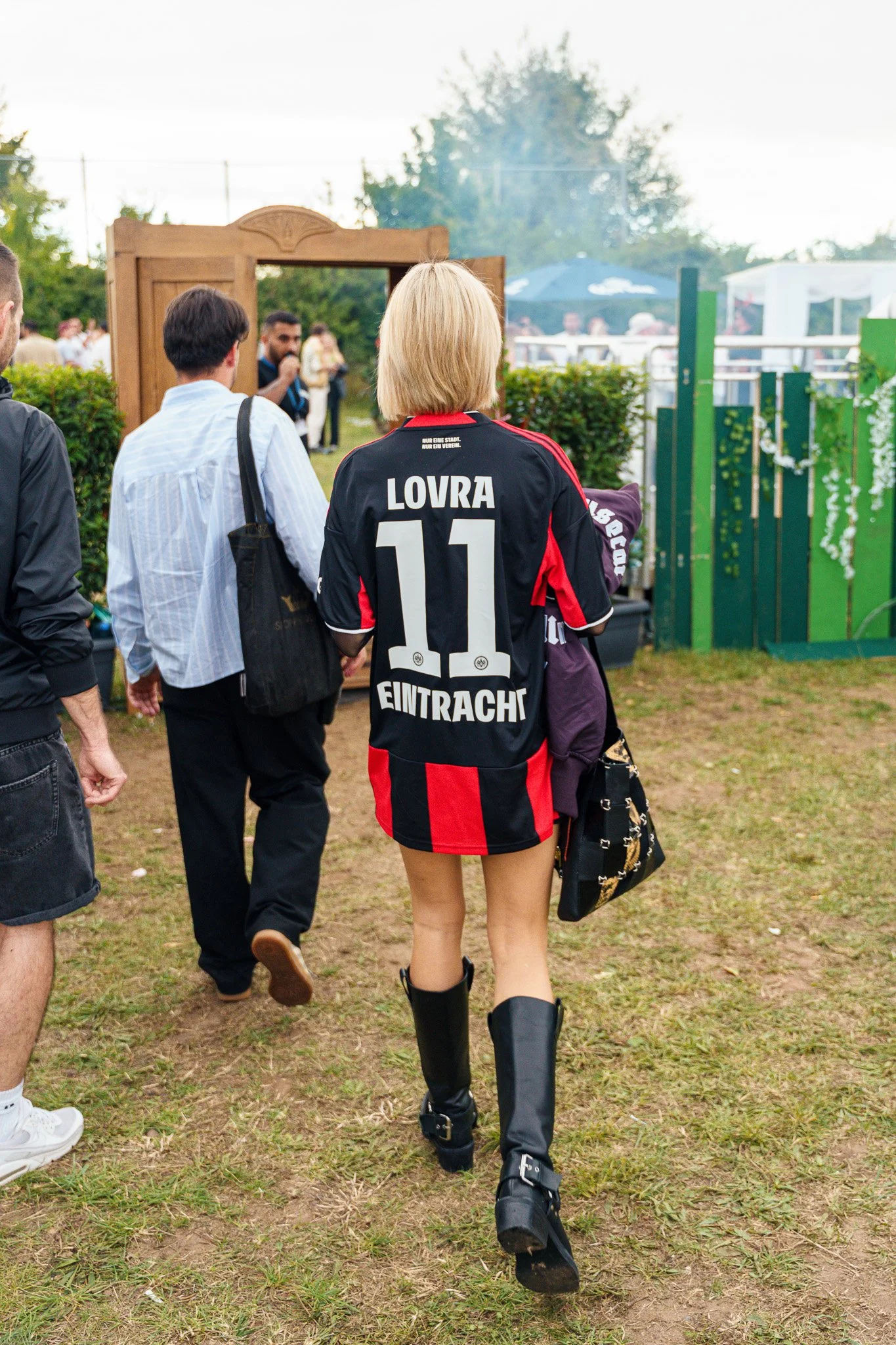 A woman wearing a black and red soccer jersey with the name "LOVRA" and the number 11, along with black boots, walking on grass at an outdoor event, with other people present.