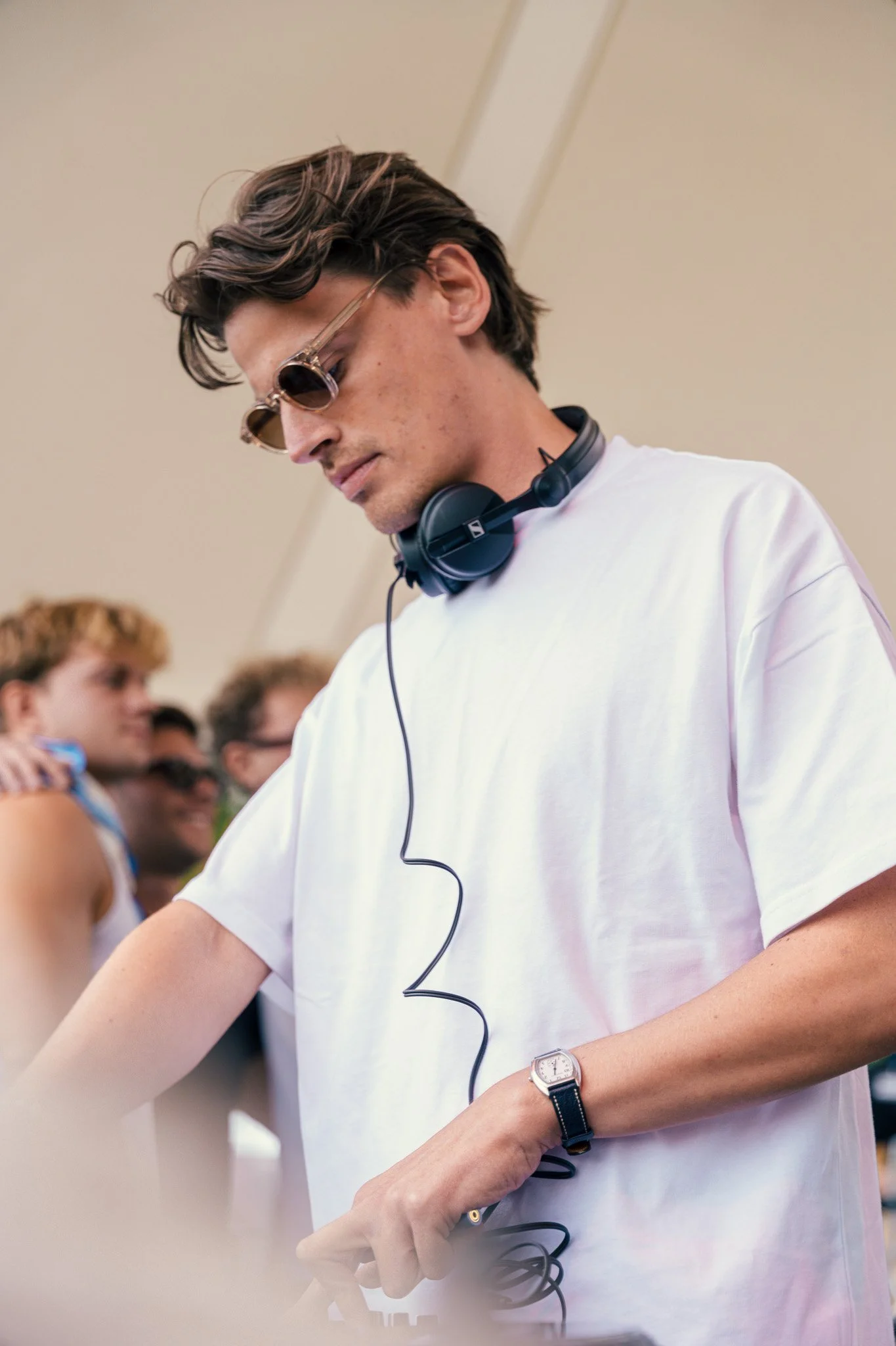 A young man DJ wearing sunglasses and a white t-shirt, focused on mixing music with headphones around his neck, while a group of people watches him in the background.