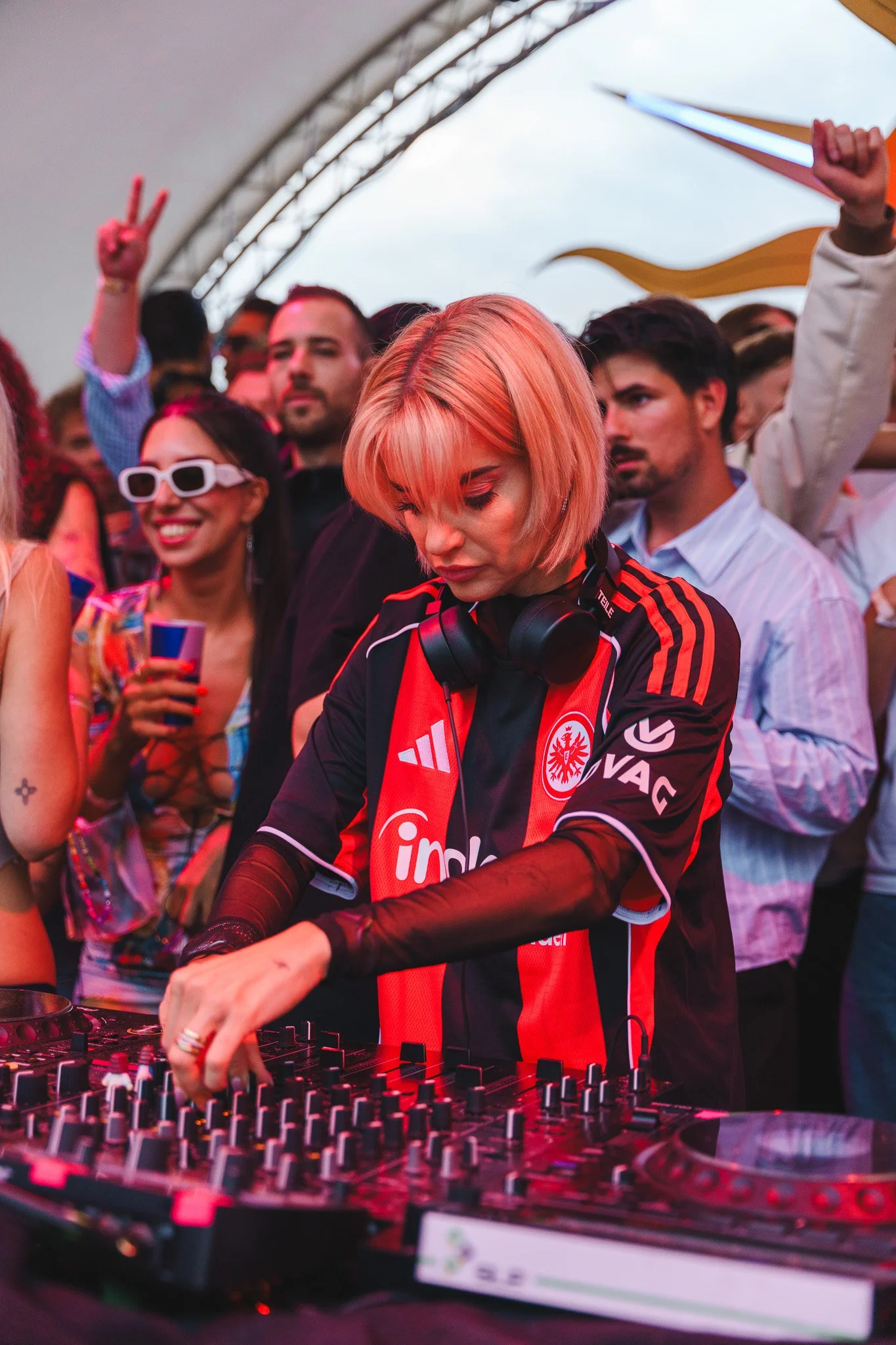 A female DJ with short blonde hair wearing a black and red soccer jersey, with headphones around her neck, mixes music at a crowded event with people dancing and enjoying the party under a tent.