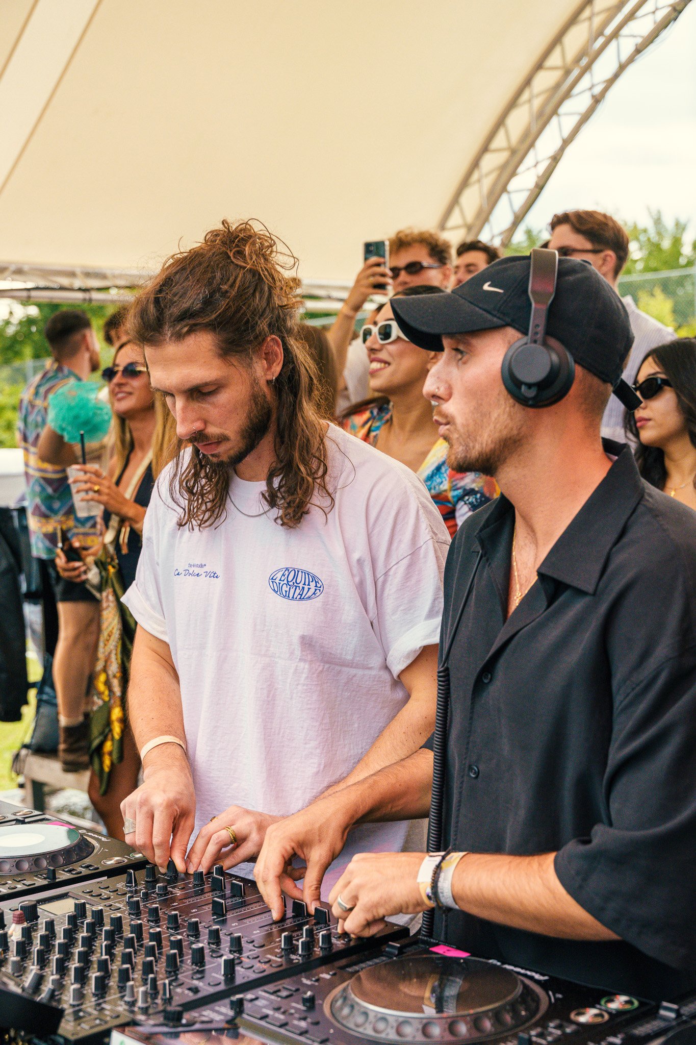 Two male DJs are mixing music on a DJ controller at an outdoor event, surrounded by a diverse crowd of people enjoying the party.