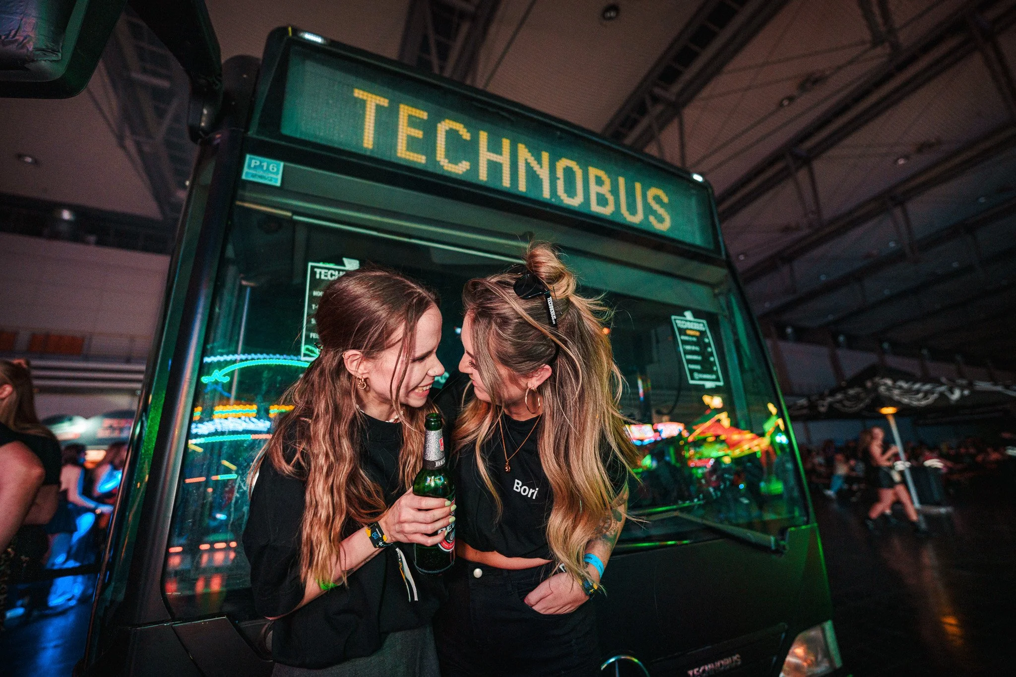 Two women smiling and leaning close together near a green TechnoBus at a festival or event, one holding a beer bottle, colorful lights in the background.