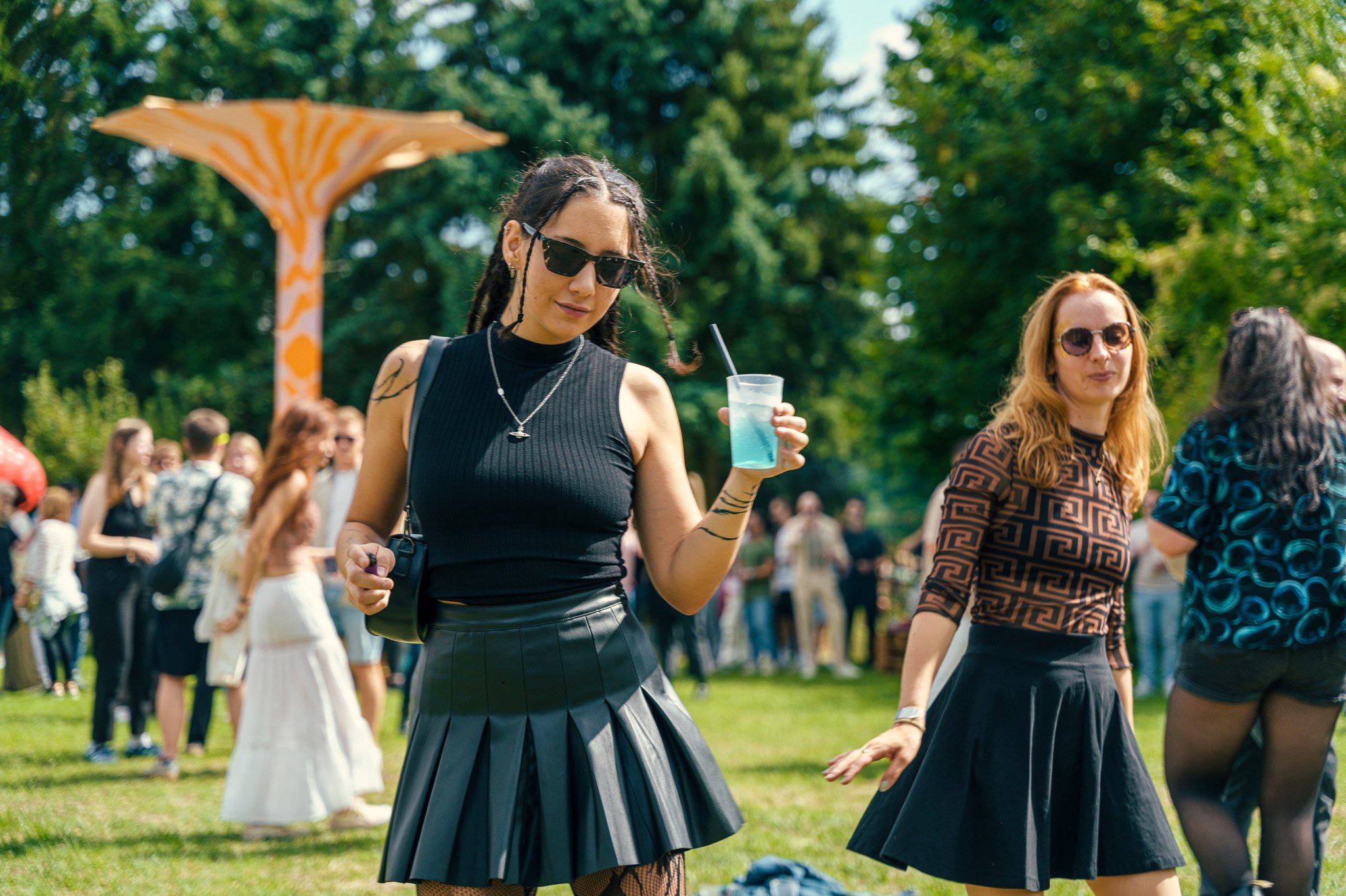 Two women dancing outdoors in a park during a sunny day, with one holding a blue cocktail.