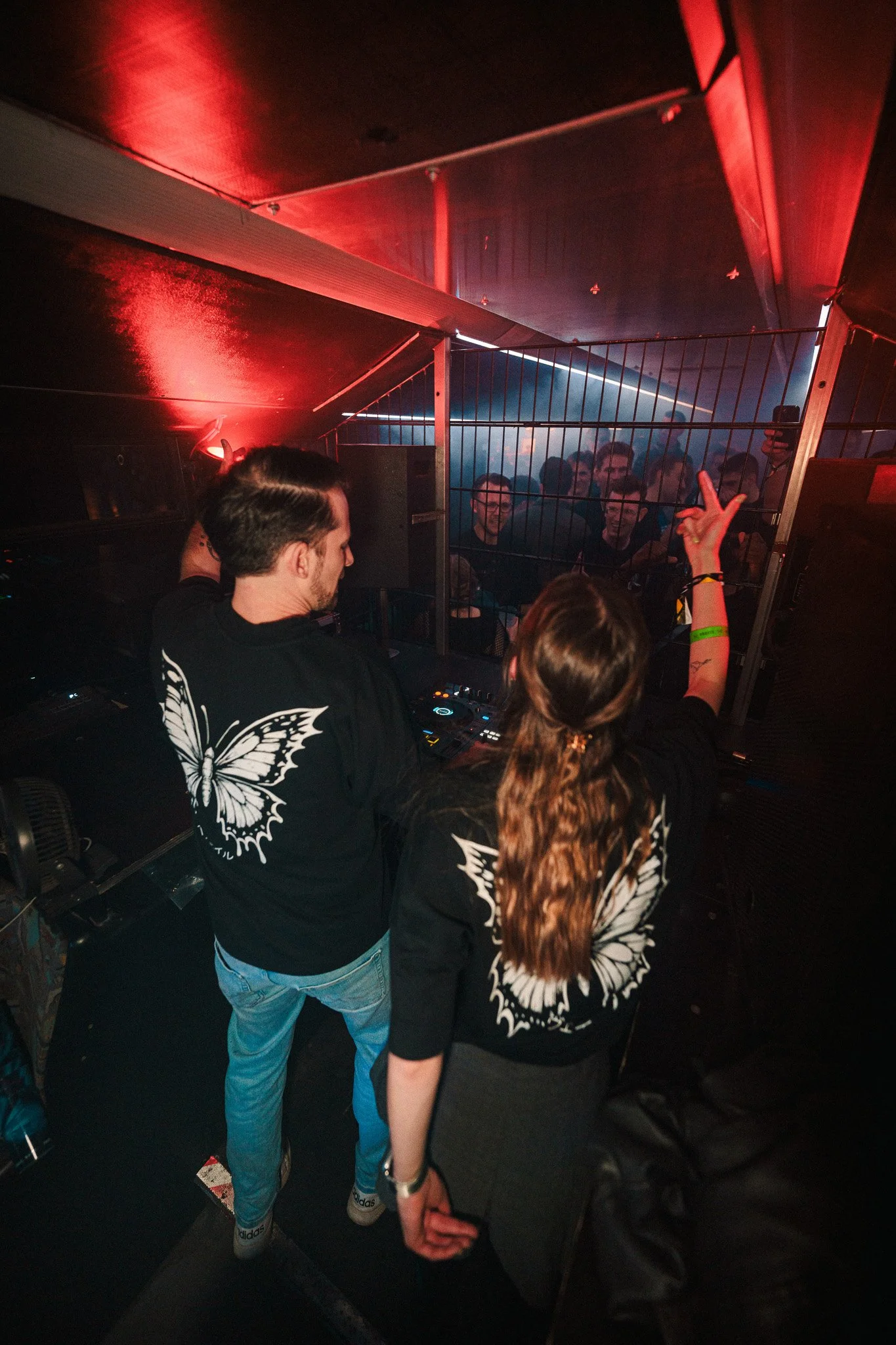 DJ and a woman with butterfly graphic on their shirts performing at a nightclub with dancing crowd behind a metal barrier, illuminated by red and blue lights.