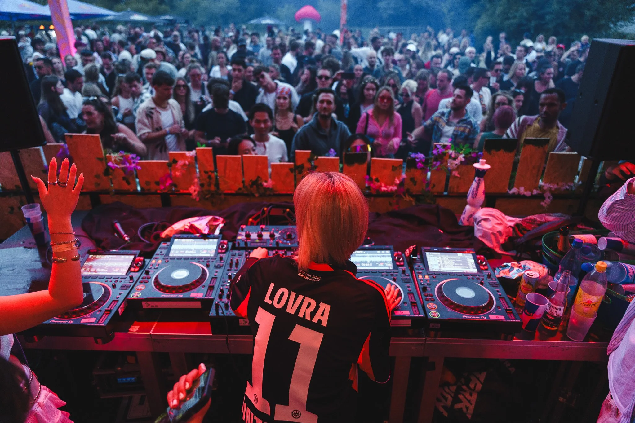 Female DJ with blonde hair playing music at a crowded outdoor event, seen from behind, with the crowd dancing and enjoying the concert.