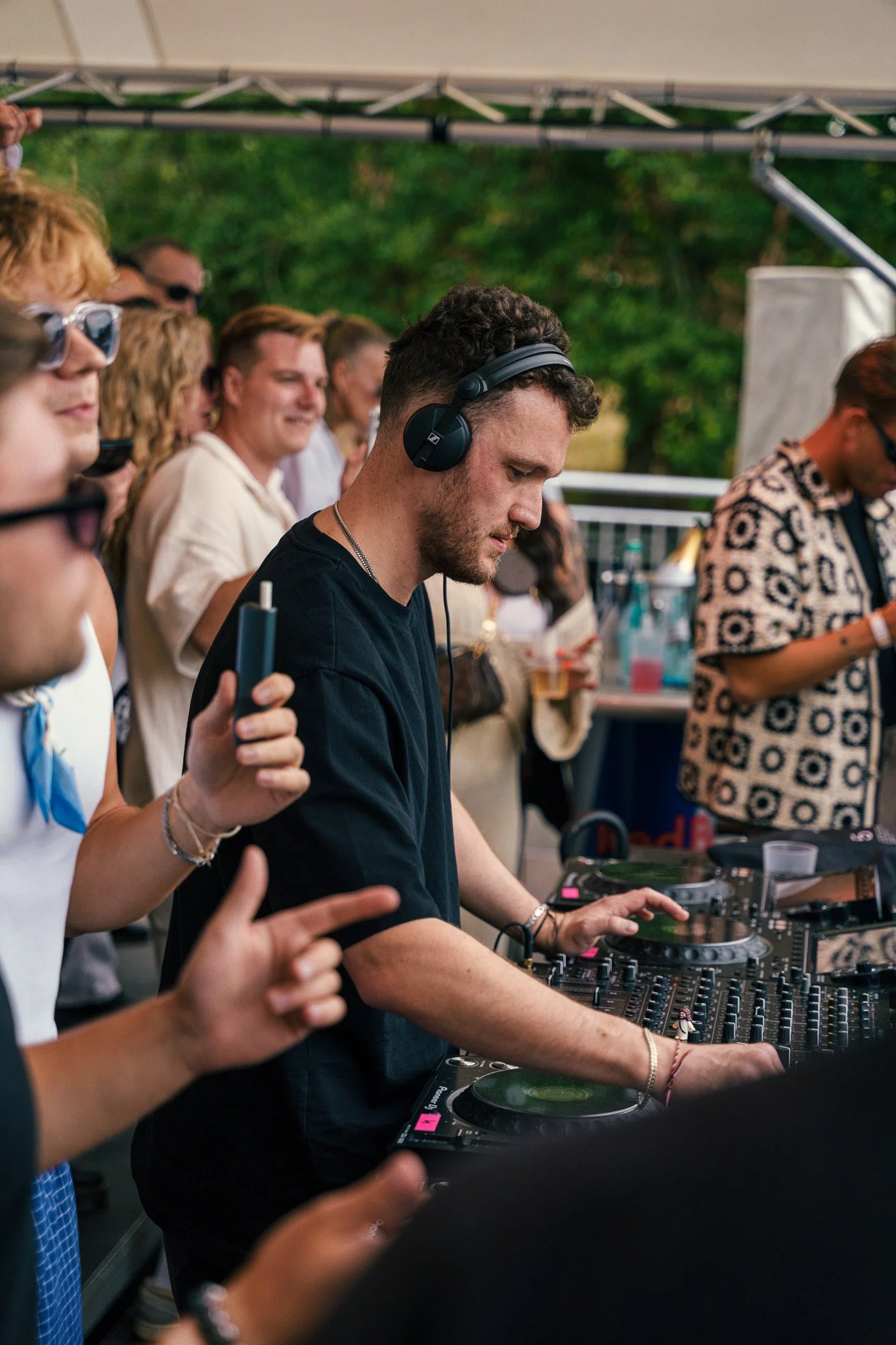 A man DJing at an outdoor event, surrounded by a crowd of people. The man is wearing headphones and focused on the DJ mixer.