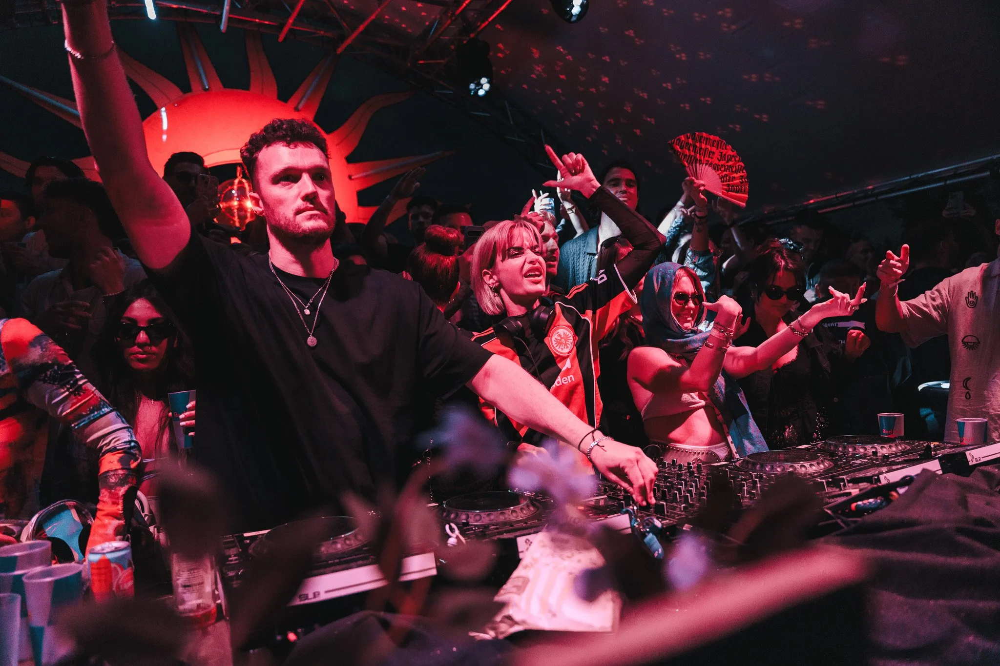 People dancing and enjoying music at a nightclub with DJ equipment in the foreground, colorful lighting, and a vibrant, energetic atmosphere.