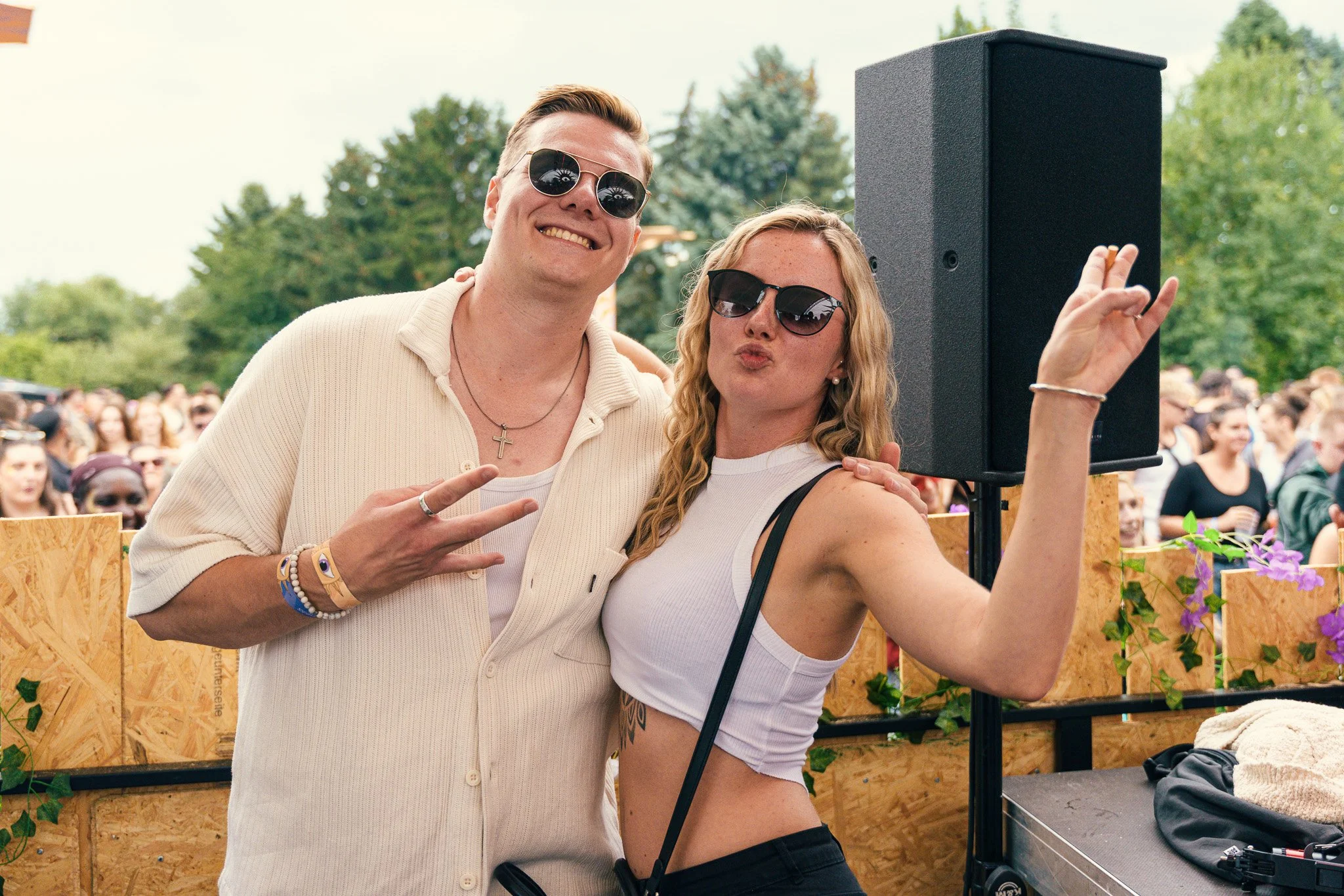 Two young adults, a man and a woman, posing together at an outdoor event during the daytime, with a crowd of people and trees in the background. They are wearing sunglasses and making peace signs, smiling and enjoying the moment.
