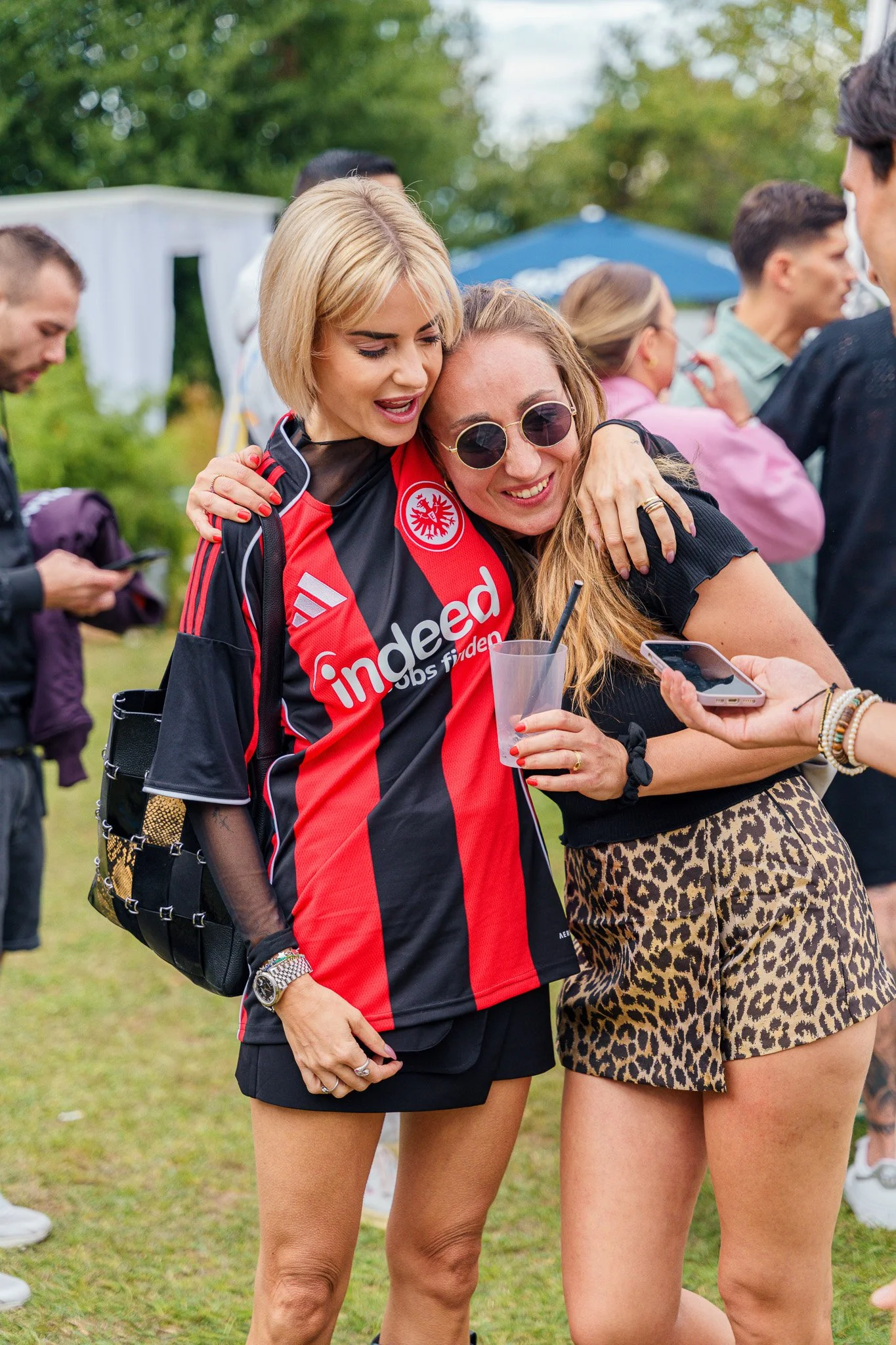 Two women hugging each other at an outdoor event, one in a red and black sports jersey, the other in a black shirt and leopard print shorts, surrounded by other people and greenery.