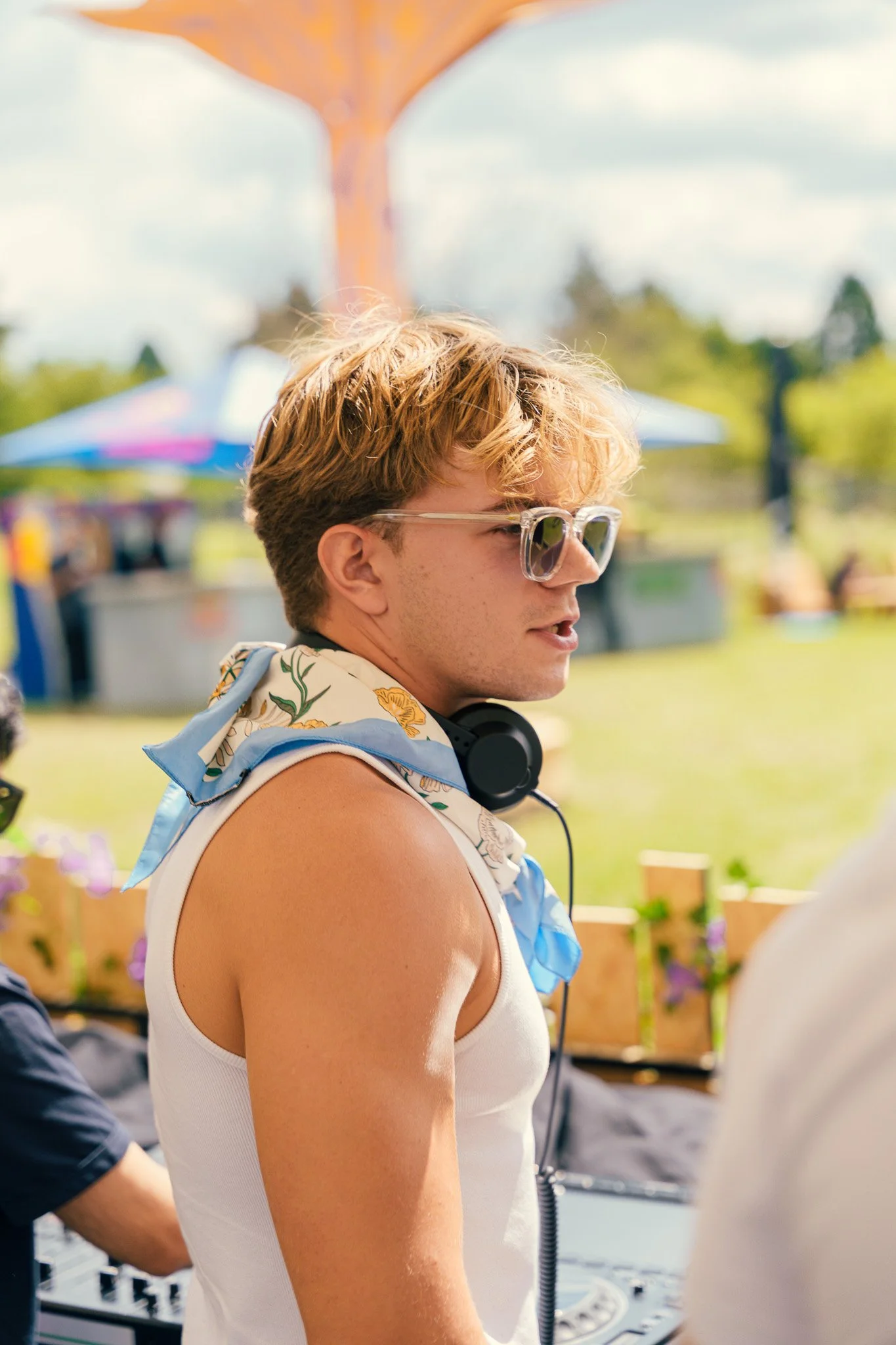 A young man with blonde hair and glasses wearing a sleeveless white shirt and a floral scarf around his neck, standing outdoors at an event with DJ equipment and a blurred background of trees and umbrellas.