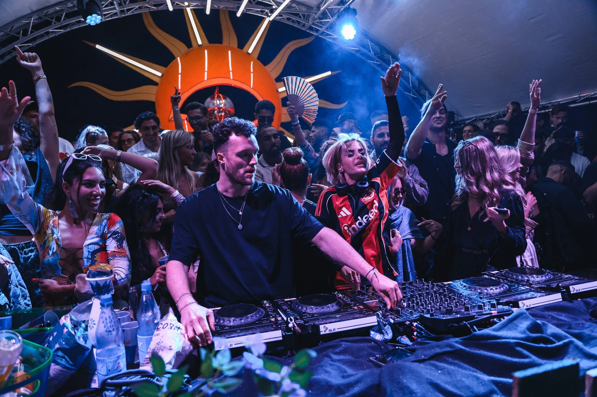 A crowded DJ booth with two DJs performing at a lively party. The scene is colorful with a sun-shaped decoration and vibrant lighting illuminating the crowd.