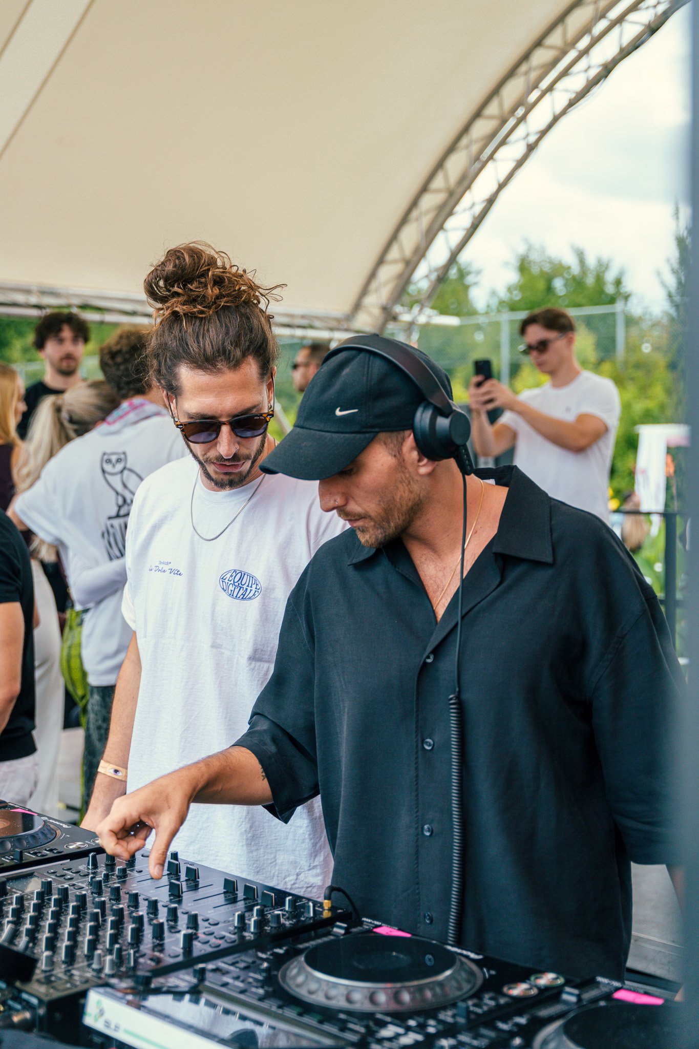 A DJ in black clothing and a black cap using a DJ controller at an outdoor event with people around.