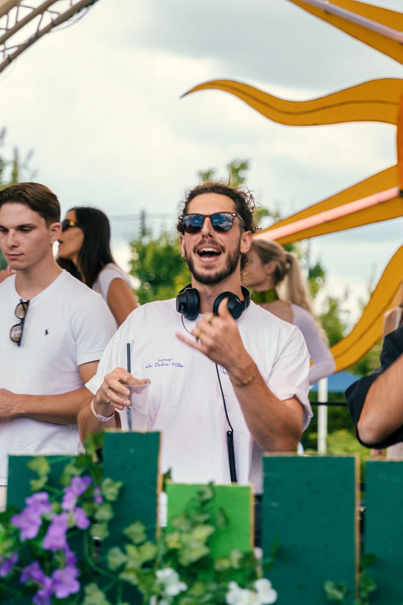 A man with sunglasses and headphones around his neck is holding a drink and smiling at an outdoor event, with other people in the background.
