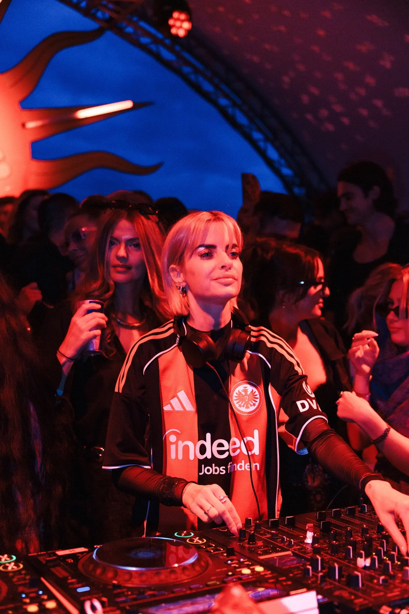 A woman DJ with short blonde hair wearing an Eintracht Frankfurt football jersey, headphones around her neck, is DJing at a nightclub or party. She is surrounded by a crowd of women and men, with a vibrant, colorful background and a large illuminated
