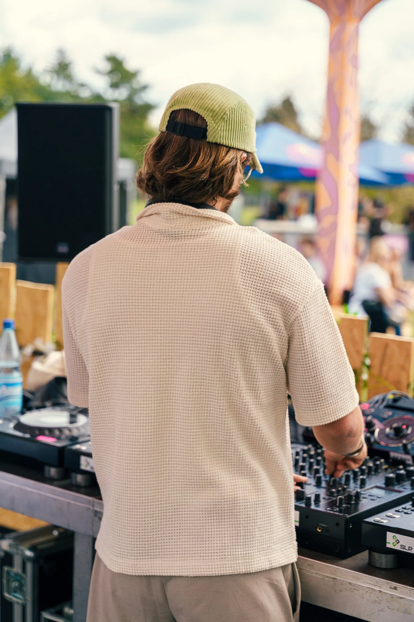 A person with shoulder-length brown hair wearing a yellow cap and beige short-sleeve shirt, operating DJ equipment at an outdoor event with tents and trees in the background.