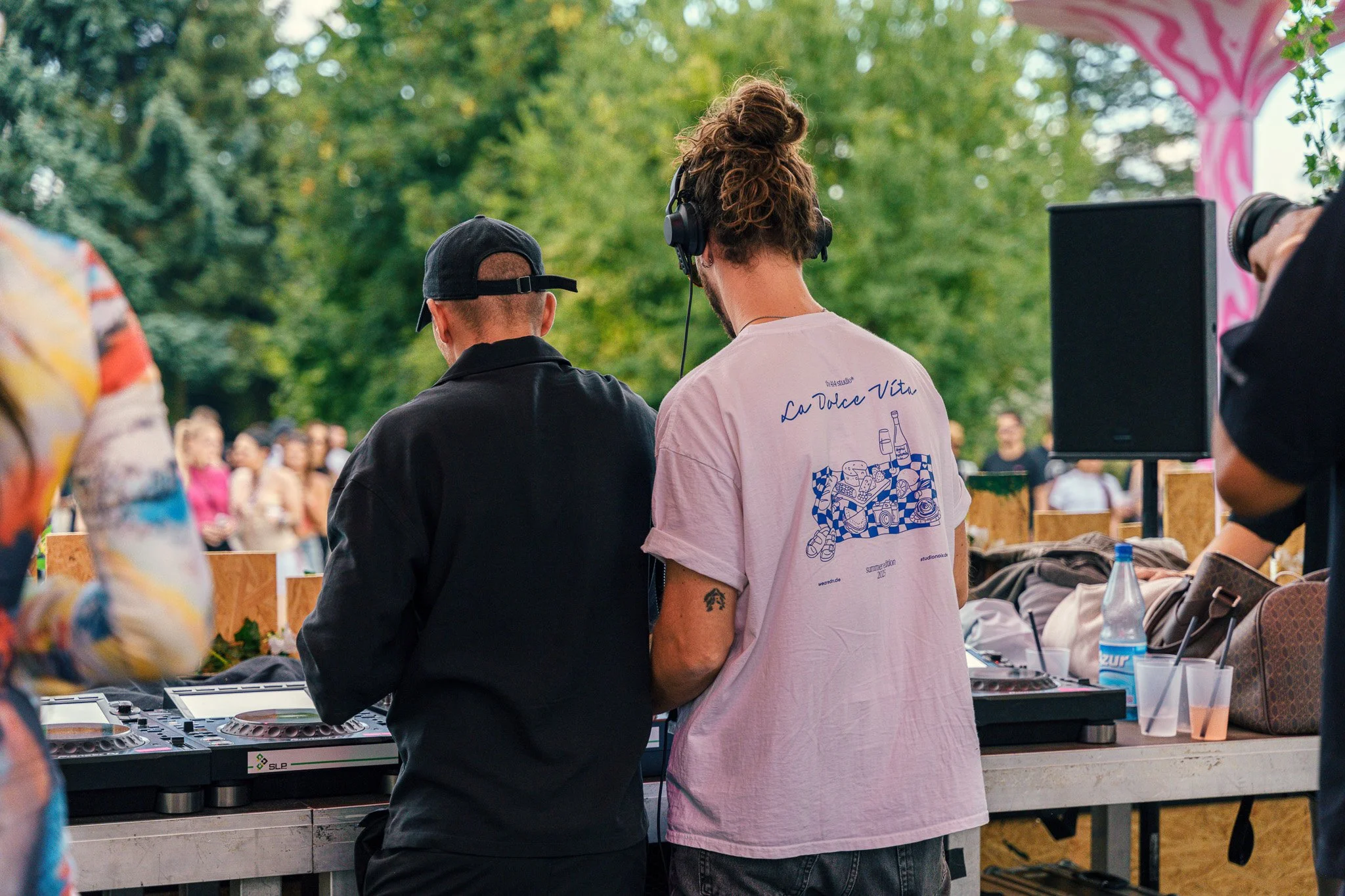 Two men are DJing at an outdoor event with trees and a crowd in the background. The man on the left wears a black shirt and cap, while the man on the right has headphones and a pink t-shirt with a blue graphic and the text 'La Dolce Vita'.