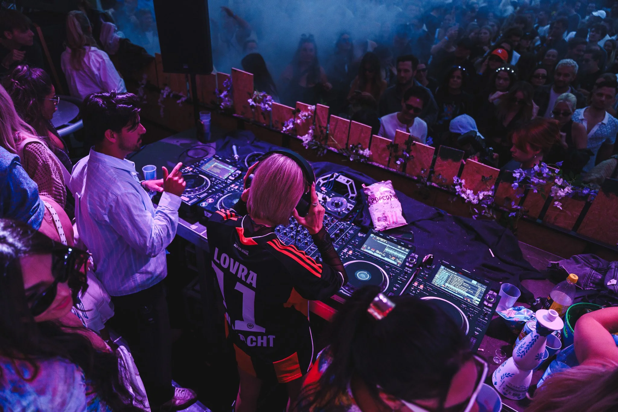 A DJ is playing music behind a crowd at a nightclub or concert, with people dancing and enjoying the event in front of the DJ booth.