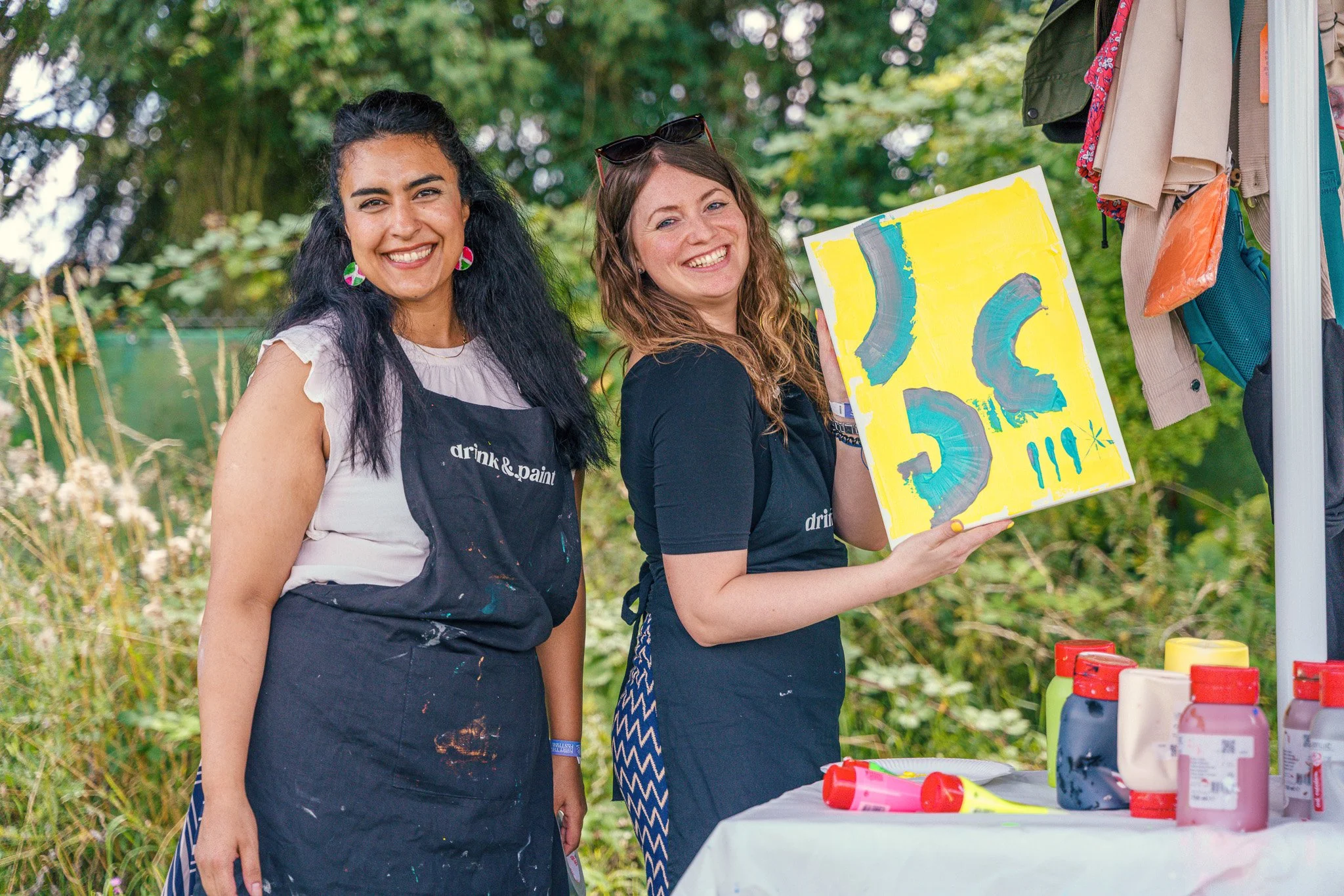 Two women smiling outdoors, wearing aprons, standing next to a table with paint bottles and supplies, one holding a colorful abstract painting.