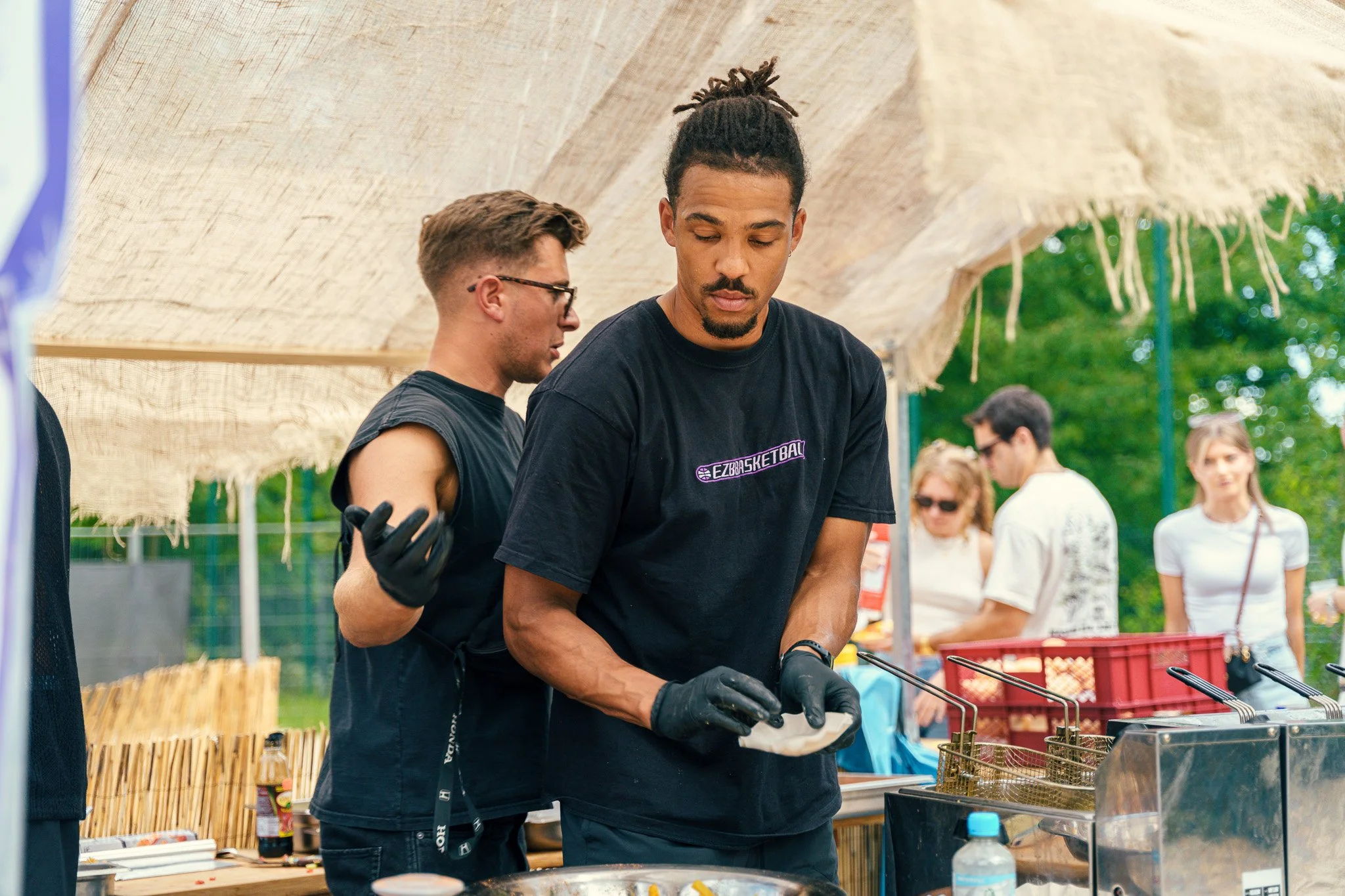 Two men preparing food at an outdoor event, with one in the foreground wearing a black T-shirt and black gloves, focused on cooking, and the other in the background wearing glasses and a sleeveless shirt, under a tent with other people and trees in t