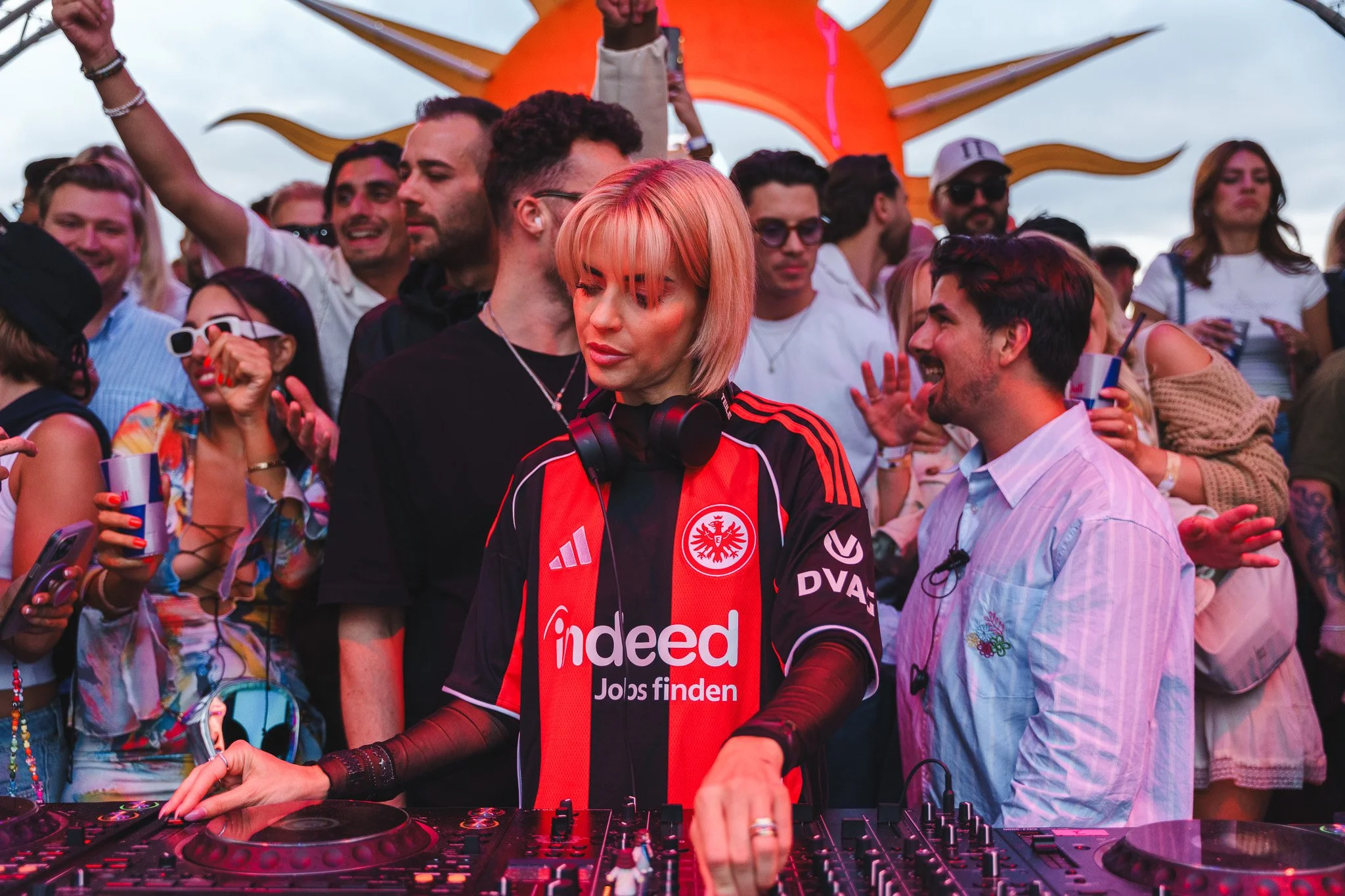 A DJ with short blonde hair wearing a red and black jersey with 'Indeed' logo, and headphones around her neck, performs at a crowded outdoor music event, with people dancing and enjoying the music in the background.