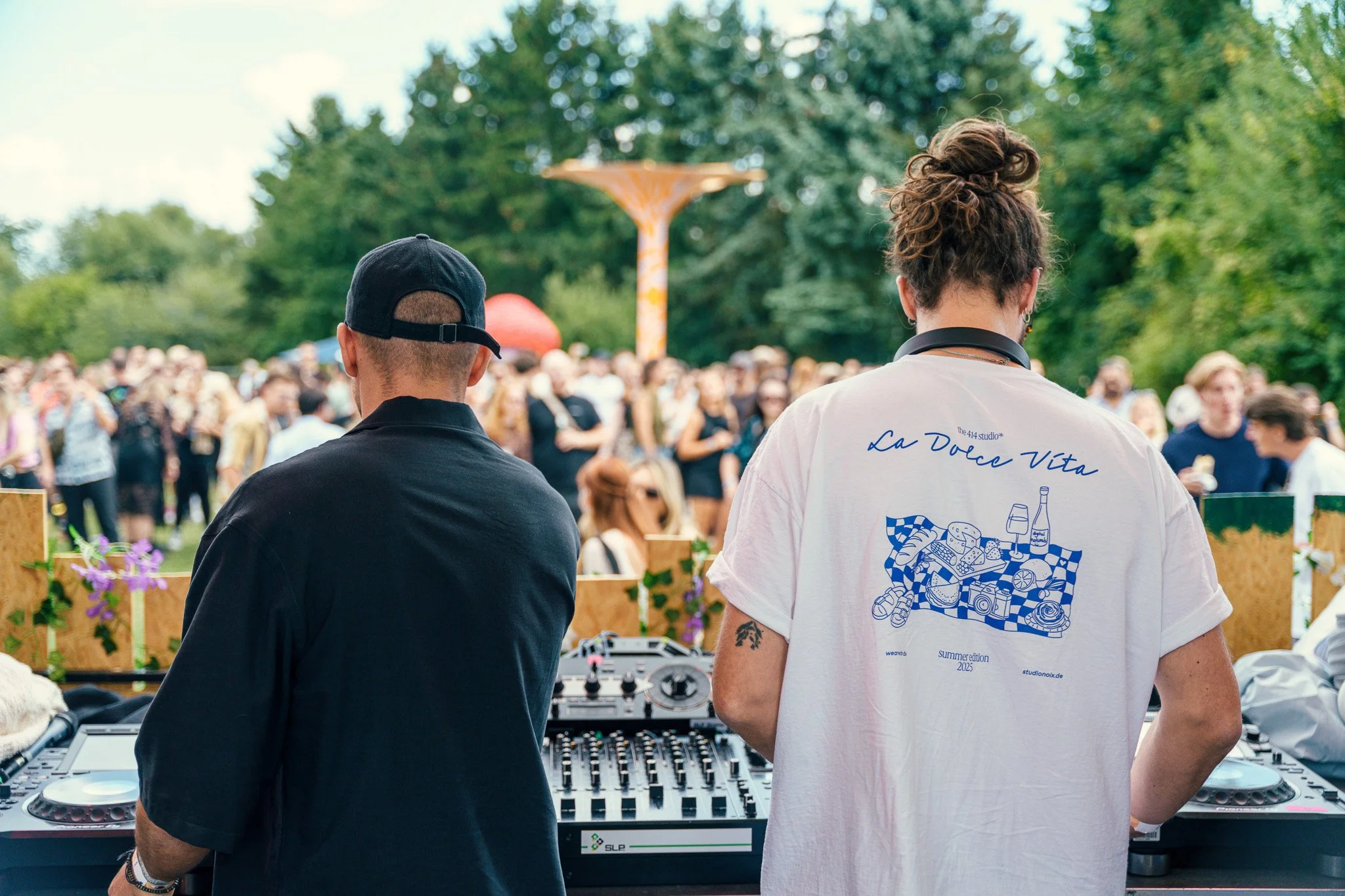 Two DJs perform at an outdoor event with a crowd of people enjoying. The DJ on the left wears a black shirt and cap, and the DJ on the right wears a white t-shirt with blue writing and illustrations, and has curly hair tied up. There is DJ equipment 